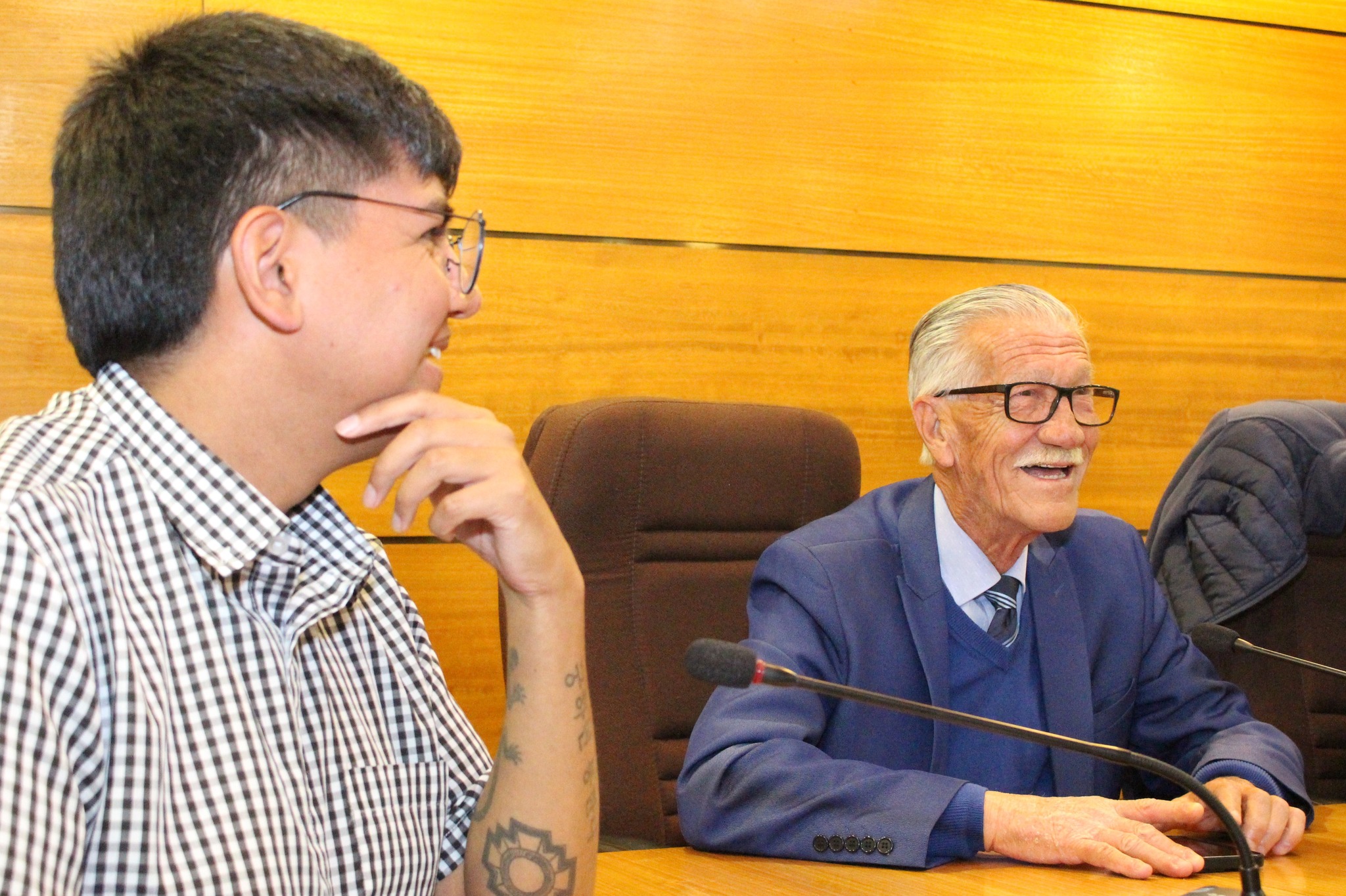 Dos asistentes durante una comisión del CORE de Antofagasta, sentados frente a micrófonos en sala de madera, mientras se exponen proyectos de Taltal.