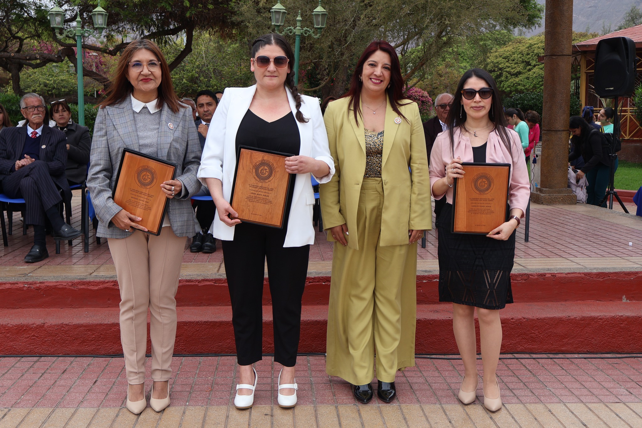 Cuatro mujeres de pie en el escenario, tres con placas de reconocimiento y una con traje mostaza; se ven asistentes y autoridades detrás en sillas.