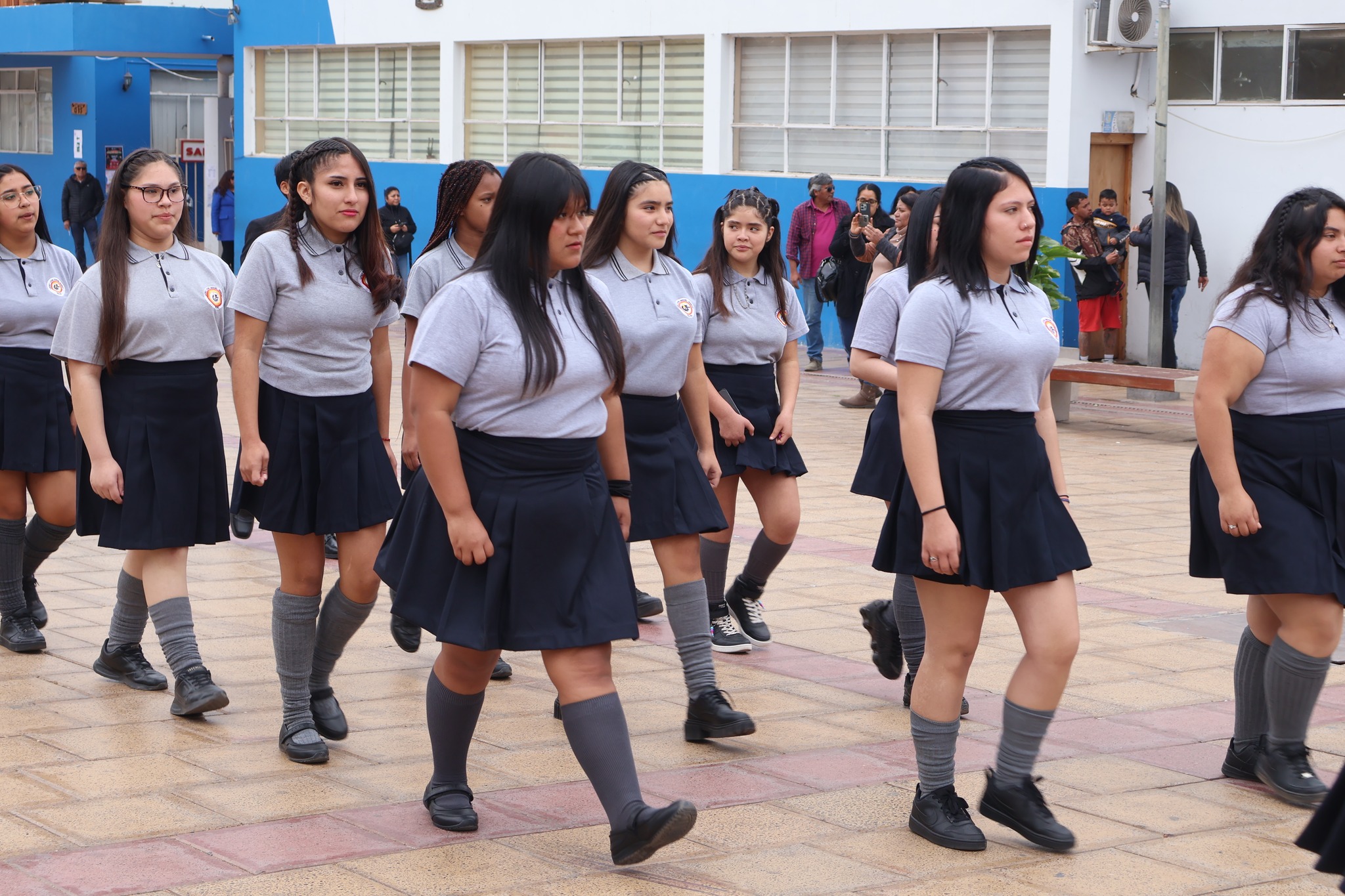 Grupo de alumnas con uniforme gris y azul marcha en formación por la plaza durante el desfile escolar.