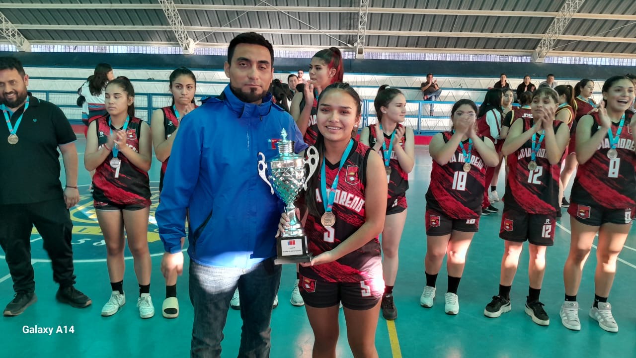 Capitana del Colegio Lidia Moreno levanta la copa de campeonas regionales; compañeras celebran en la premiación.