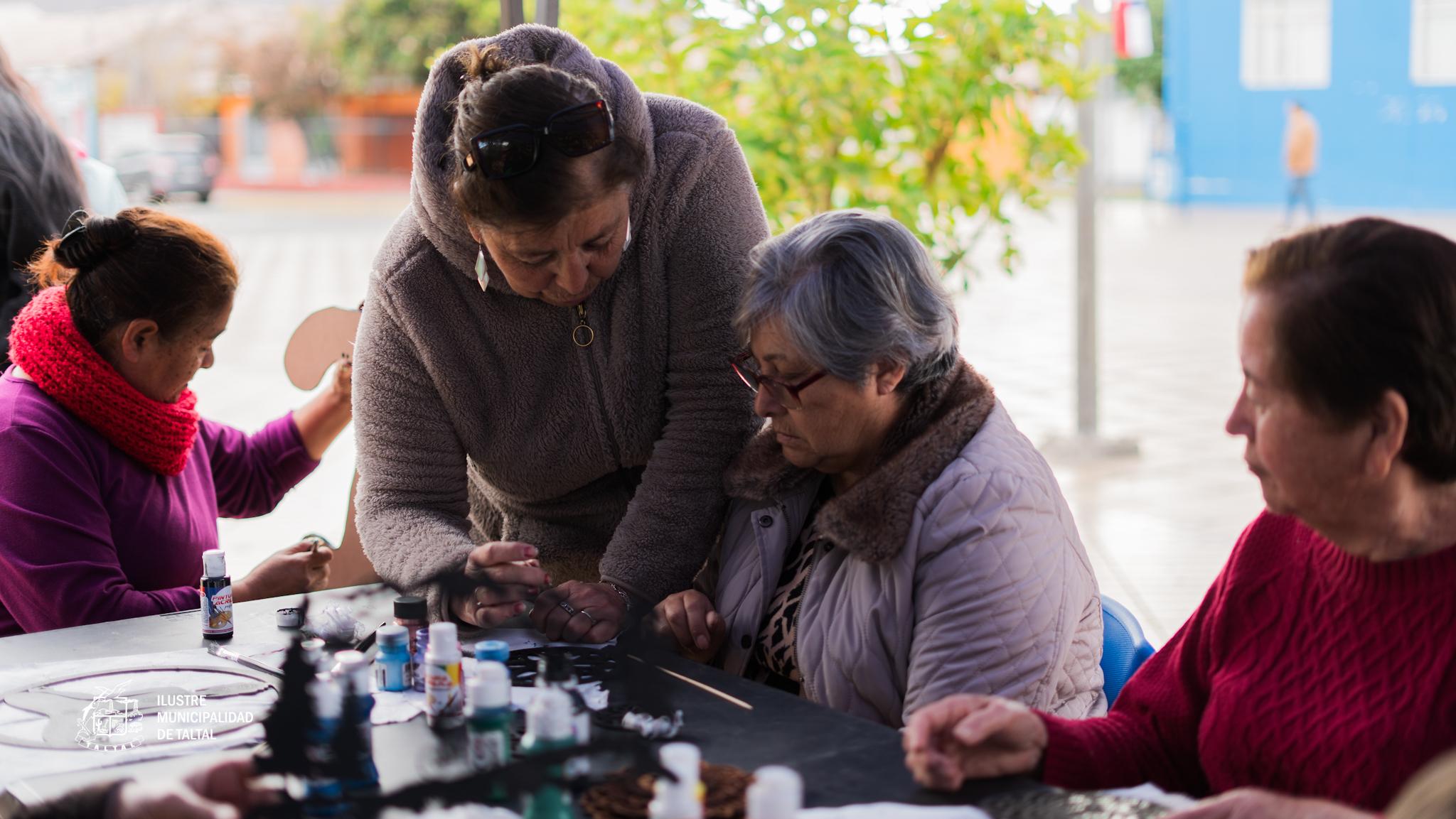 Monitora apoya a personas mayores en mesa de trabajo con pinturas y plantillas.