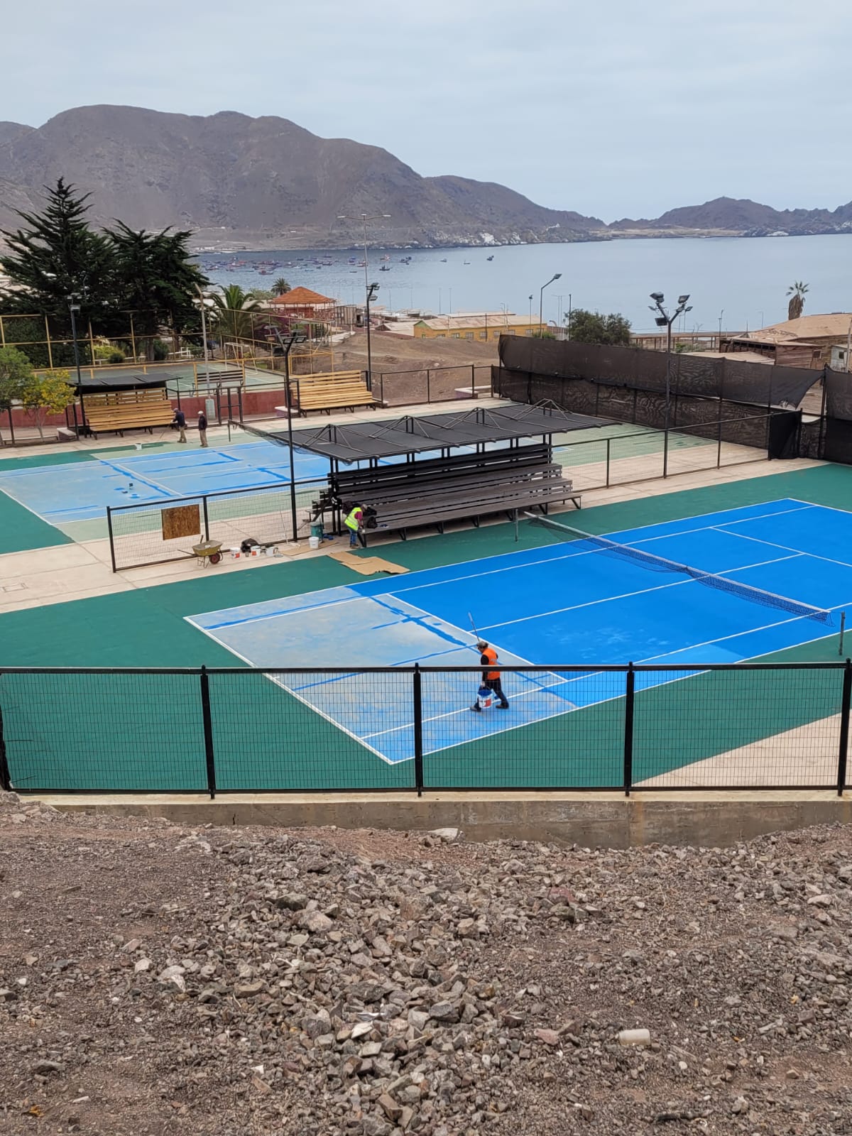 Vista en altura del complejo de canchas con graderías y mar de fondo.