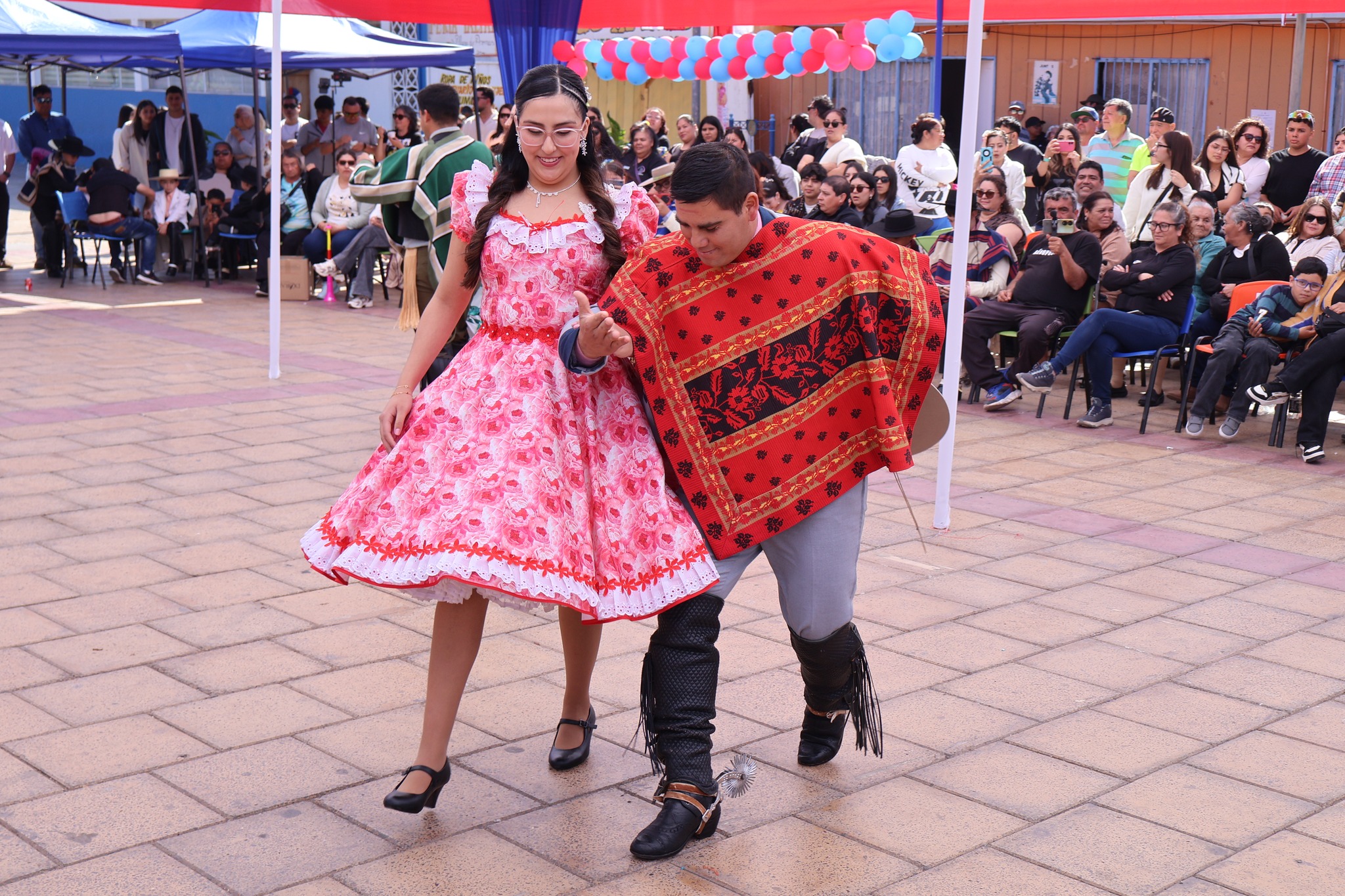 Pareja adulta compite en cueca; ella con vestido rosado florado y él con poncho rojo, pasos coordinados ante el público.