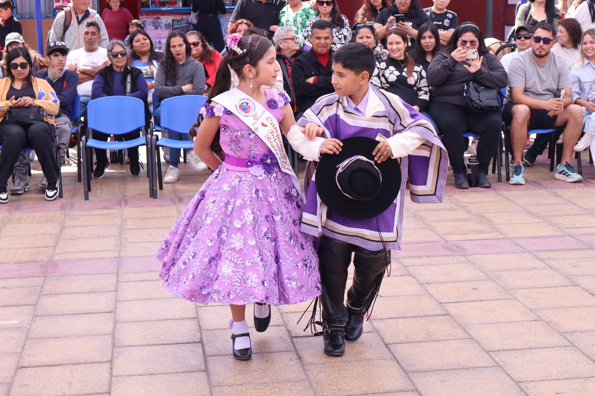 Presentación infantil destacada: pareja con trajes lilas y espuelas baila cueca frente al palco repleto.