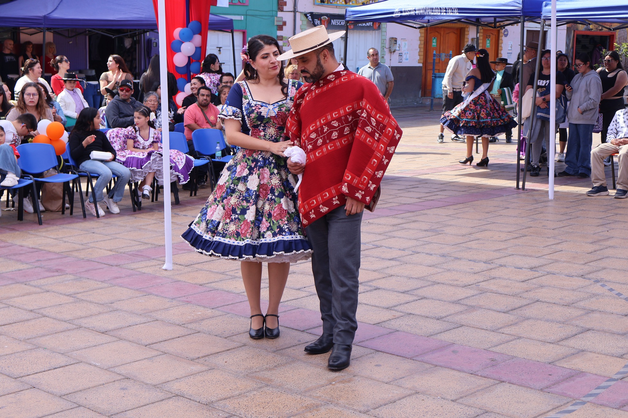 Pareja del jurado en la Plaza de Armas Arturo Prat de Taltal; él con poncho rojo y sombrero, ella con vestido florado en mano y público de fondo.