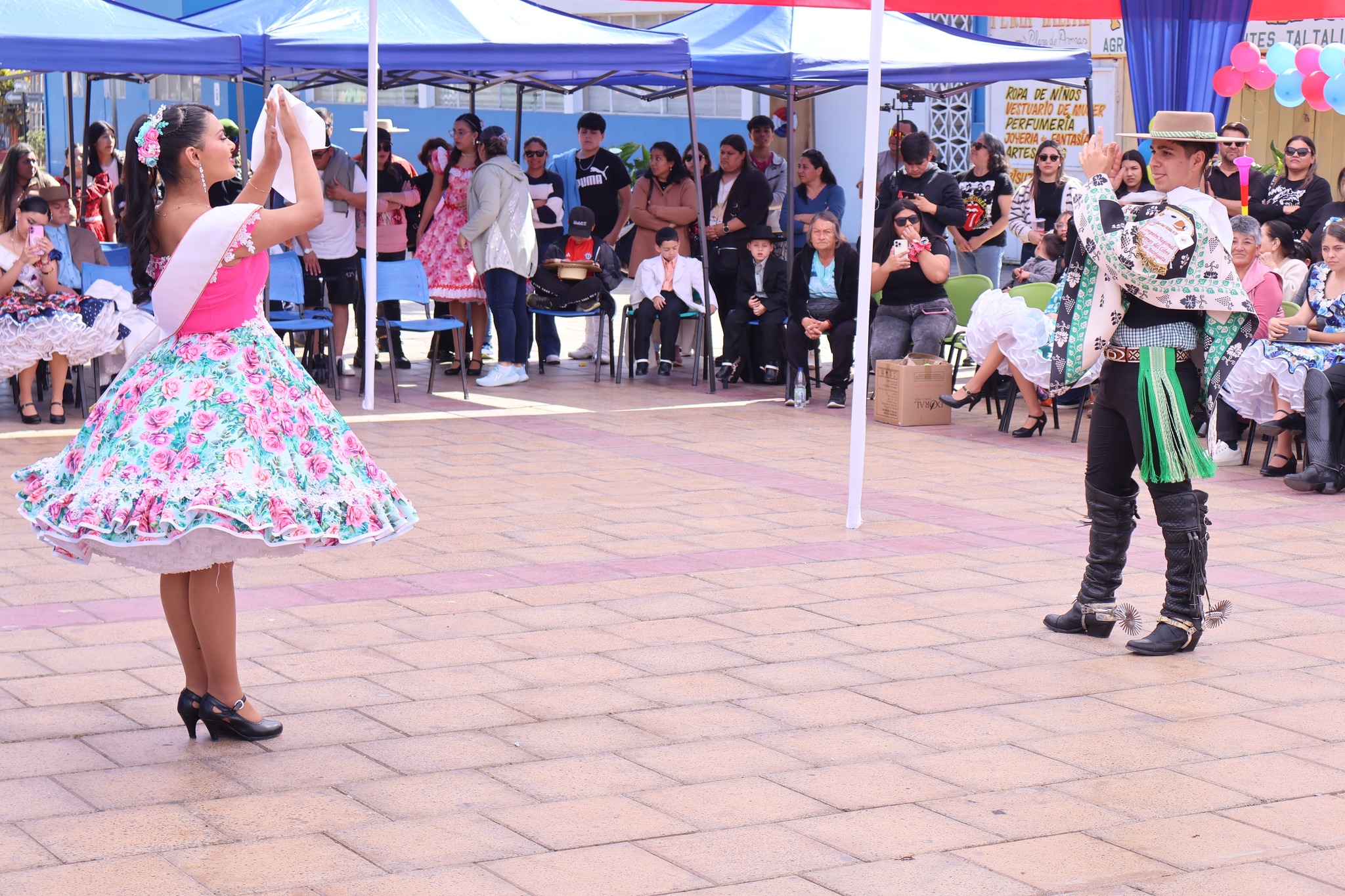 Competidores de cueca frente a frente; bailarina con vestido turquesa y banda, huaso con manta clara y espuelas, asistentes observan bajo toldos.