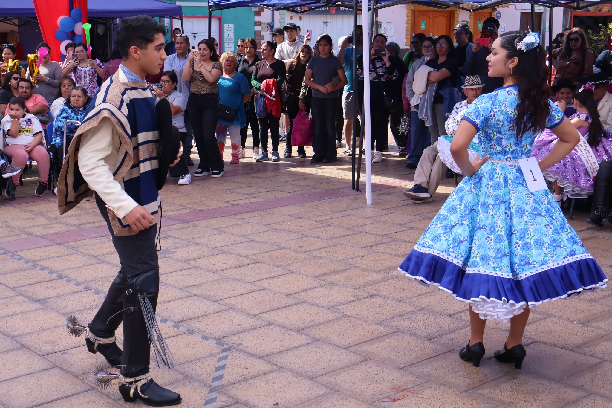 Dupla Nº1 inicia su vuelta de cueca: ella con vestido celeste y peineta, él con manta azul y botas con espuelas, rodeados de público en Taltal. 