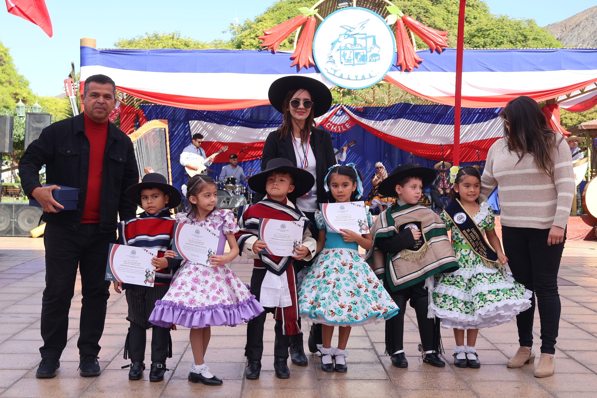 Premiación de los más pequeños: niña y niños con trajes típicos reciben diplomas junto a la organización: escenario tricolor de fondo.