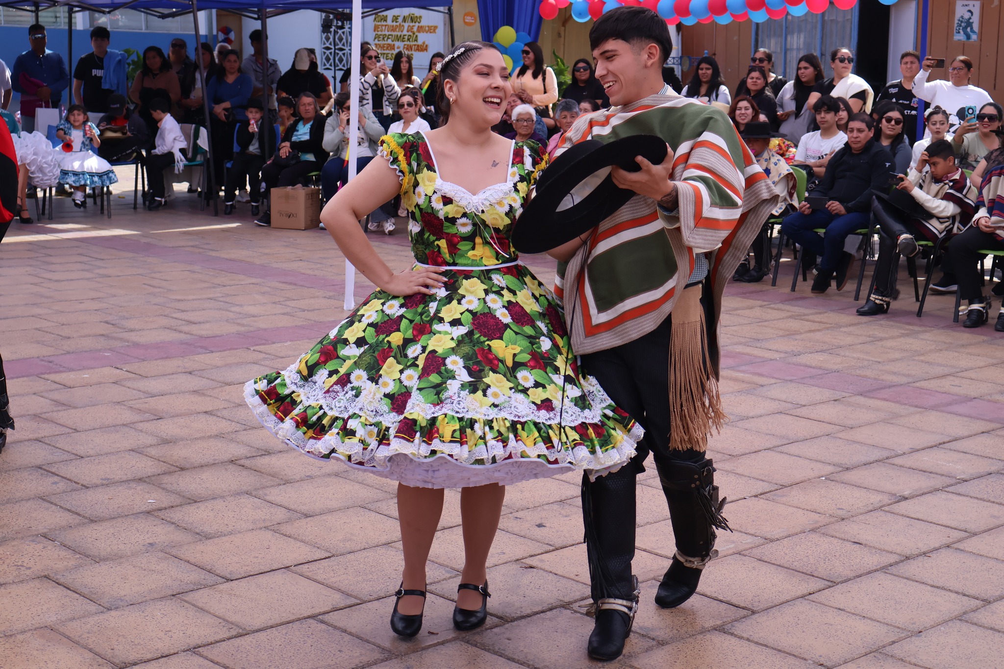 Pareja de cueca sonríe al finalizar su pie en la Plaza Arturo Prat de Taltal, ella con vestido floriado y él con manta verde y sombrero en mano, Campeonato Comunal 2025.