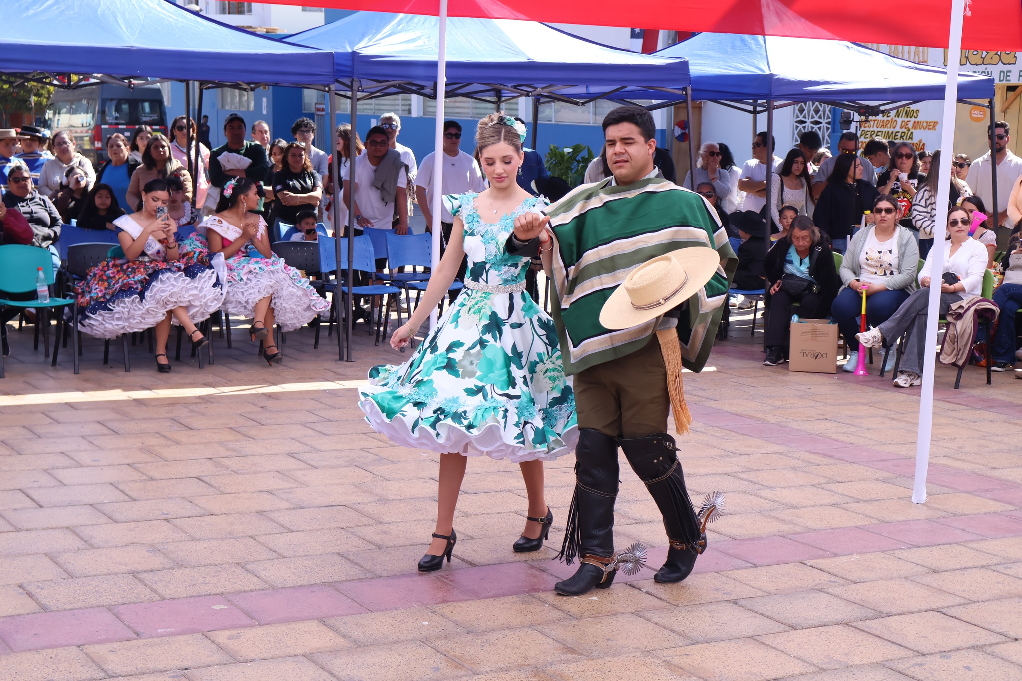 Pasada de una pareja en categoría superior: bailarina con vestido azul floral y huaso con manta verde; público aplaude alrededor.