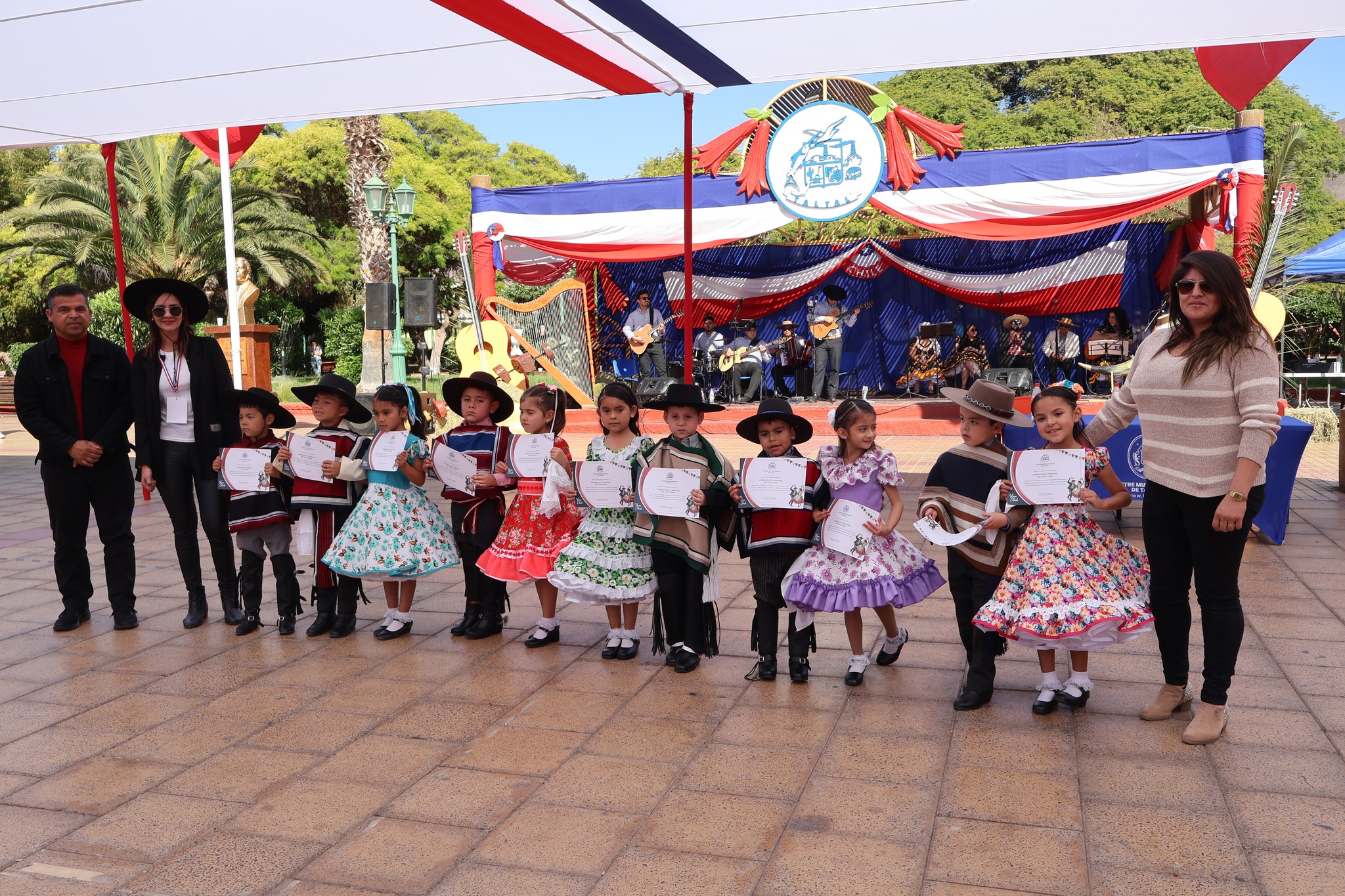 Niñas y niños con trajes típicos reciben diplomas en la premiación del Campeonato Comunal de Cueca 2025 en la Plaza Arturo Prat de Taltal, junto a autoridades y músicos en el escenario.