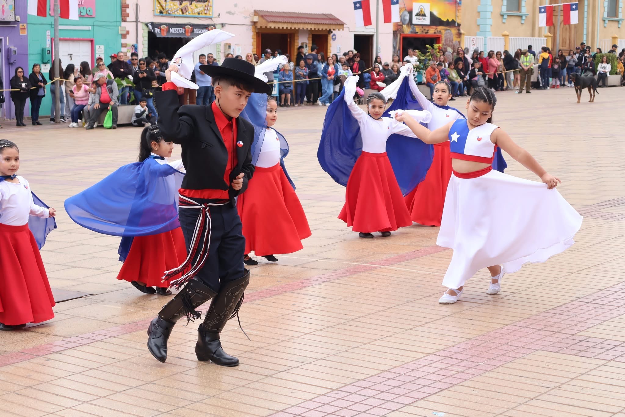 Pareja infantil de huaso y dama ejecuta figura de cueca, con elenco de apoyo al fondo.