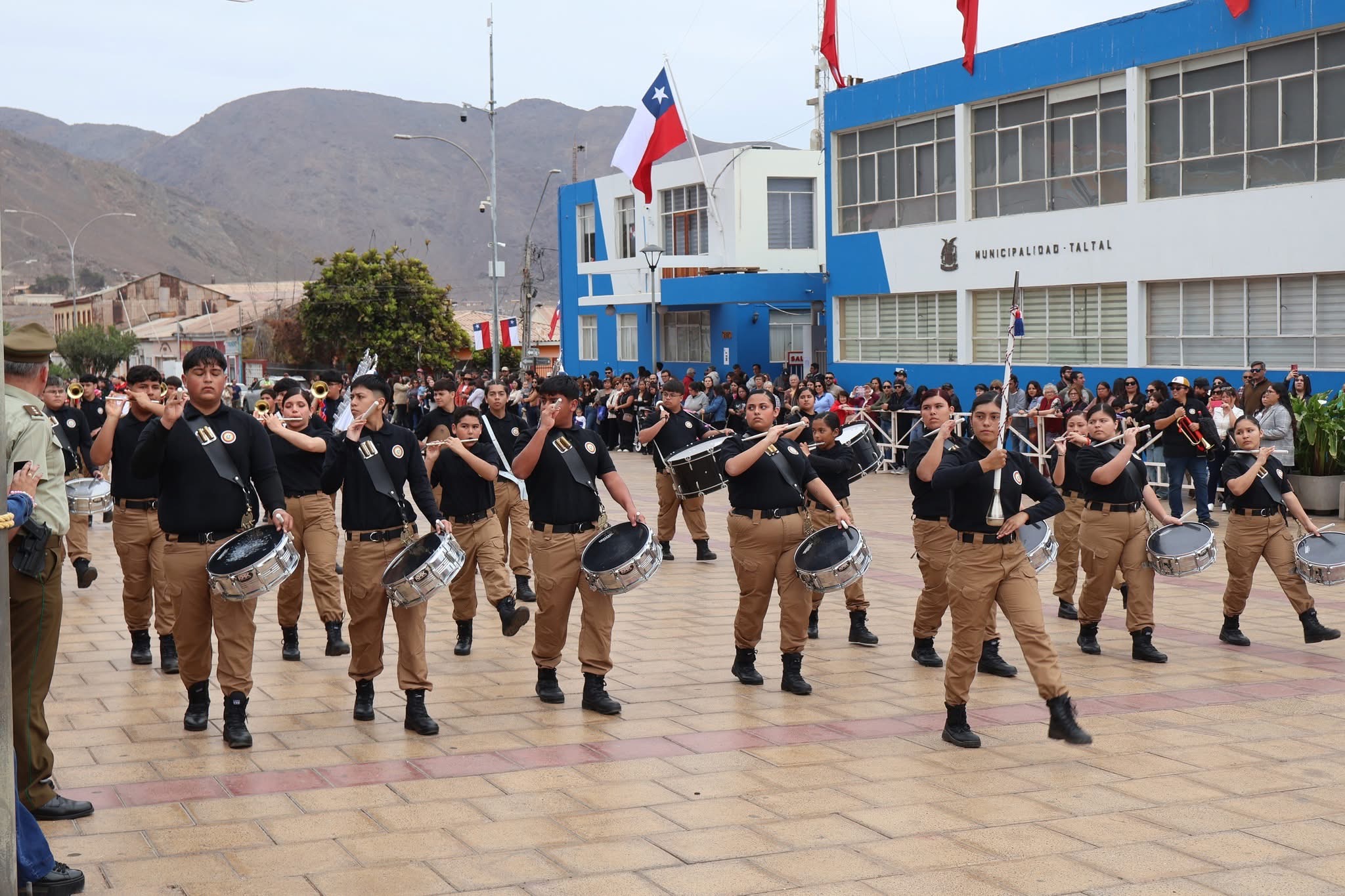 Banda de guerra desfila frente al edificio municipal ante gran marco de público.