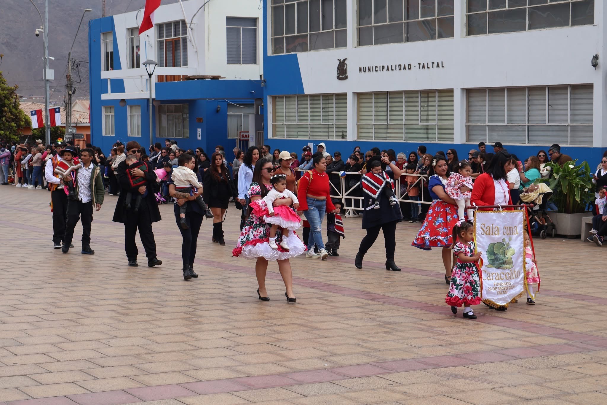 Sala cuna Caracolito participa en el desfile; familias y lactante avanzan con vestimenta dieciochera por la Plaza de Armas de Taltal.