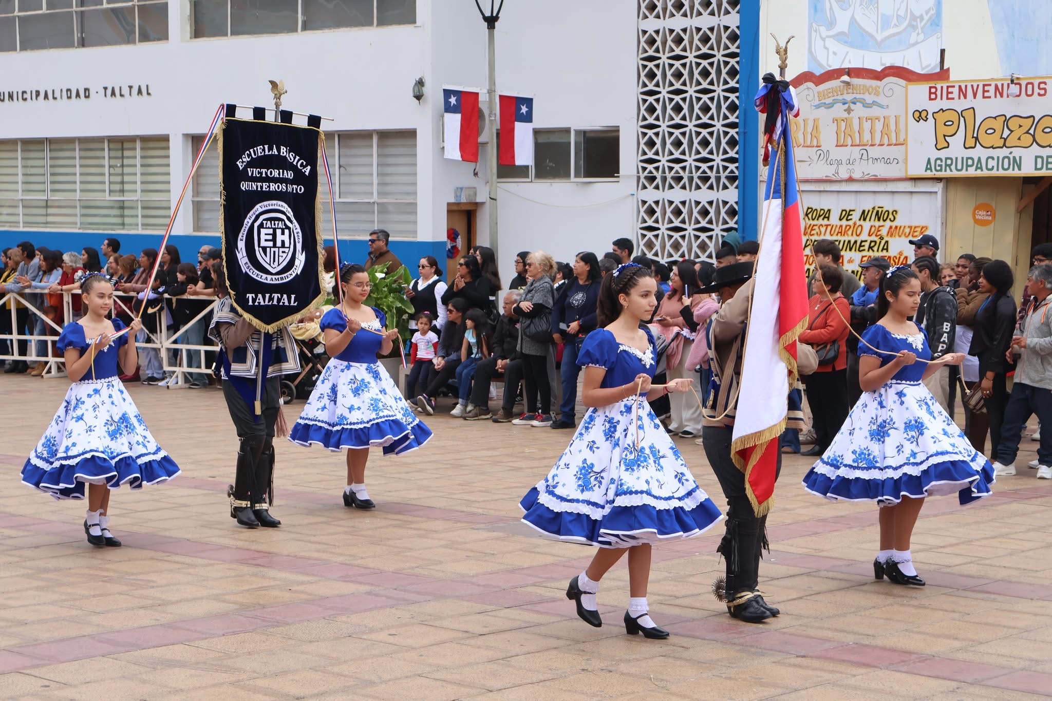 Alumnas con vestidos azules y blancos escoltan el estandarte y la bandera en el desfile escolar de Taltal.