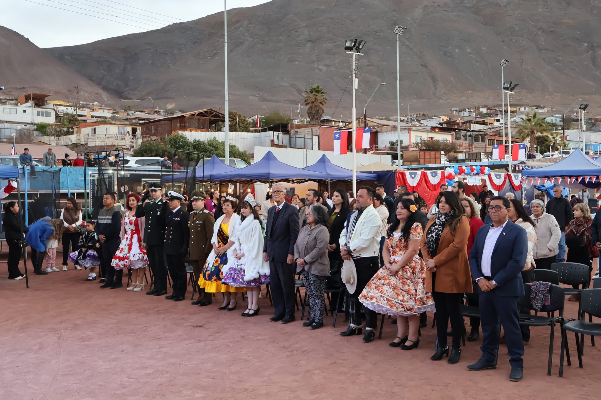 Acto protocolar de inauguración con autoridades, invitados y vecinos de pie frente a los stands y banderines dieciocheros, al pie de los cerros de Taltal.