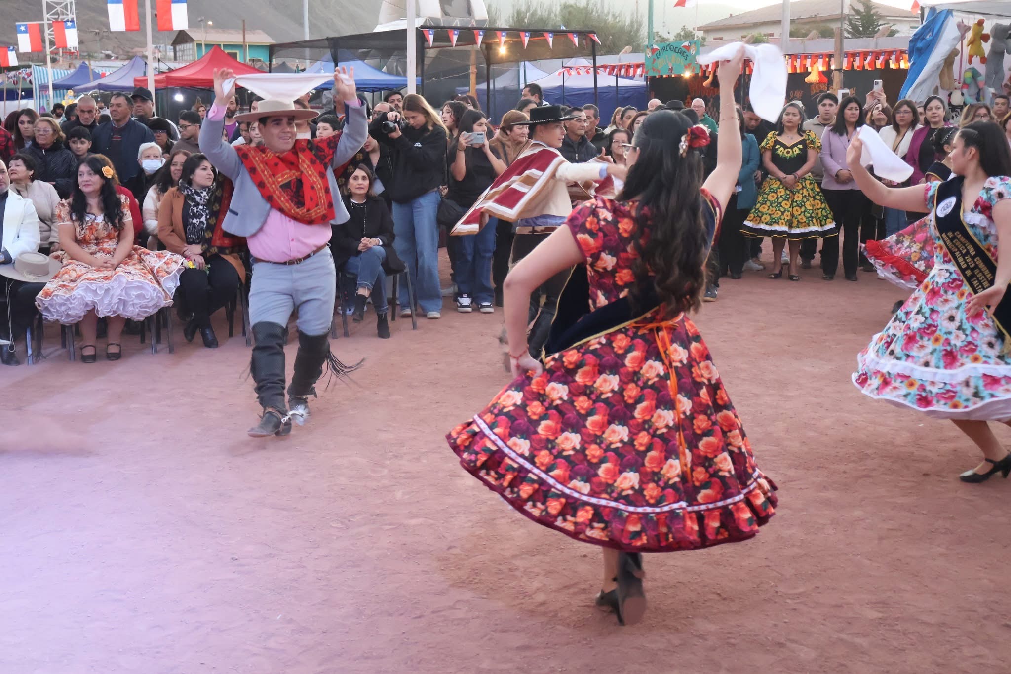 Pareja infantil en traje tradicional -niña con vestido floreado y niño con manta y sombrero- interpretan cueca entre juegos típicos y público.