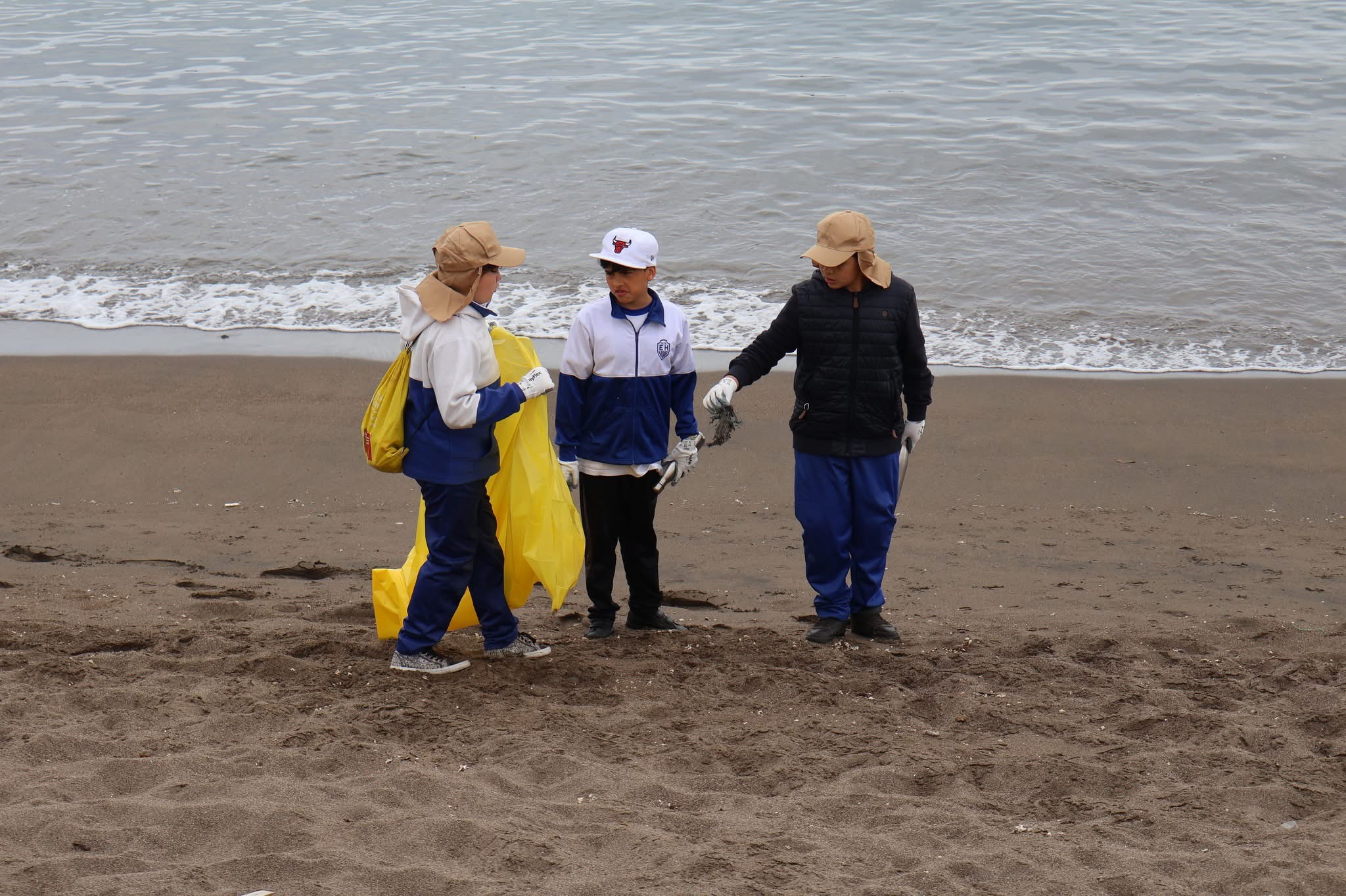 Niños con uniformes escolares y bolsas amarillas participando en la limpieza de la playa en Taltal.