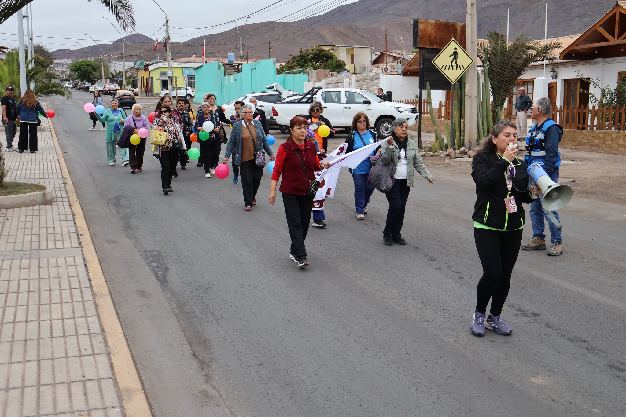 Desfile de adultos mayores en la calle, con una organizadora hablando por megáfono.