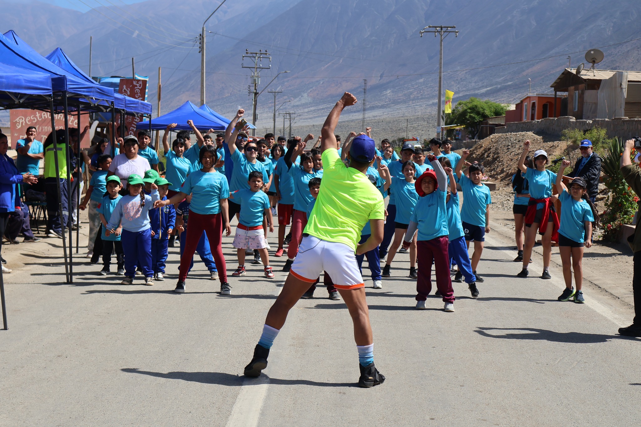 Instructor lidera el calentamiento masivo con decenas de vecinos antes de la Corrida Familiar de Paposo 2025, con fondo de cerros y carpas municipales.