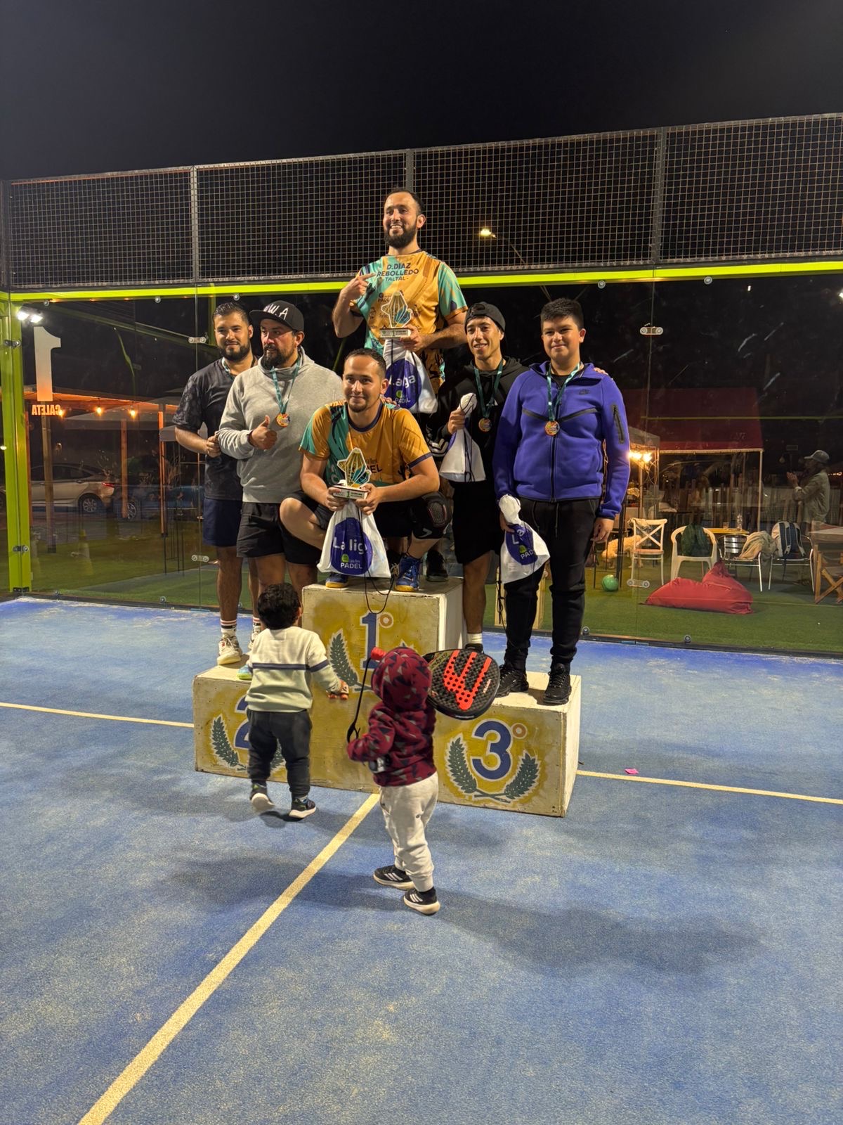 Jugadores de 5ta categoría varones celebrando en el podio con trofeos, medallas y apoyo familiar durante la premiación de La Liga Caleta Hueso Pádel 2025.