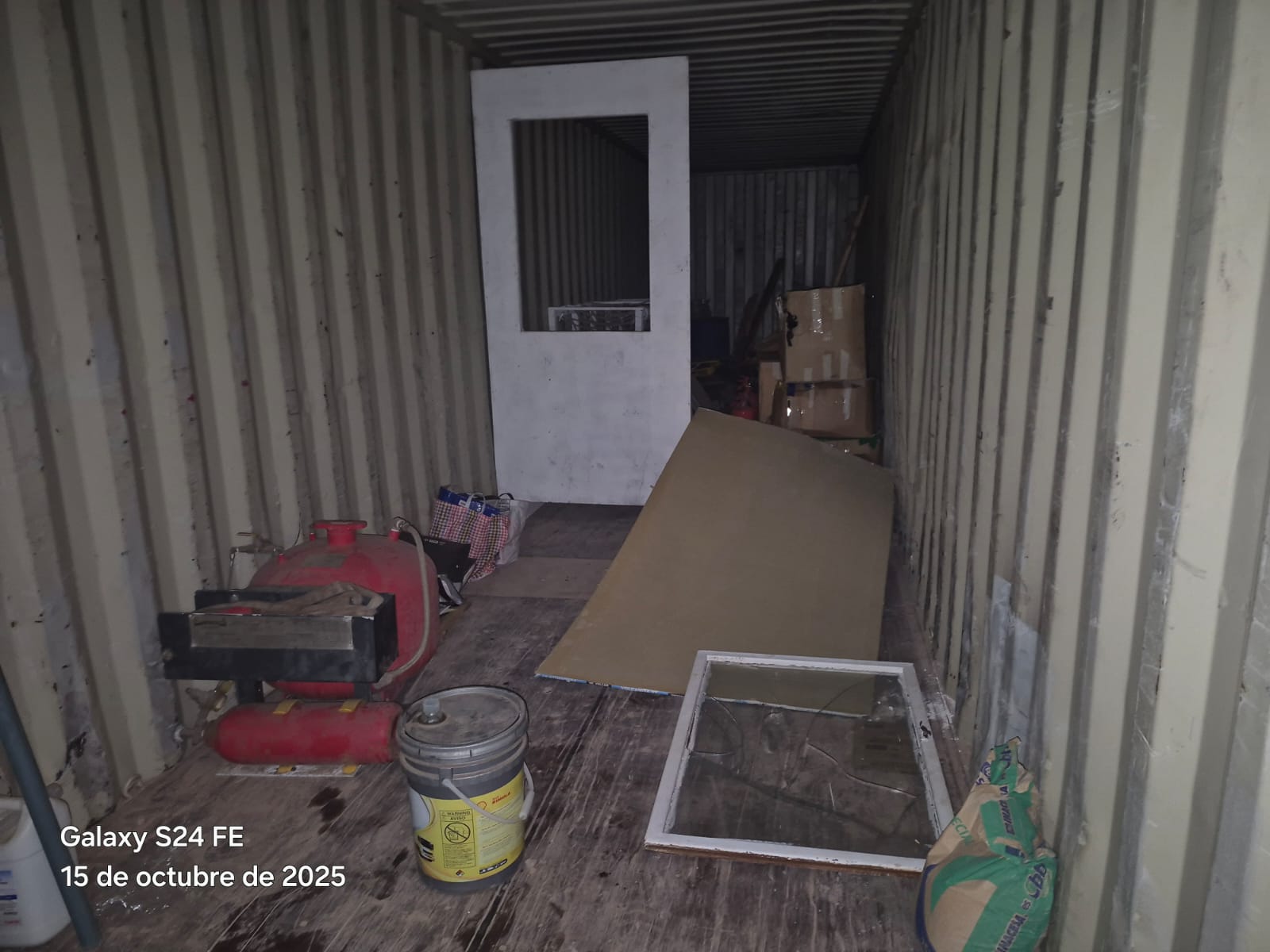 Interior de un contenedor con materiales de construcción desordenados, incluyendo una puerta blanca, un compresor rojo, cubetas y sacos de cemento.