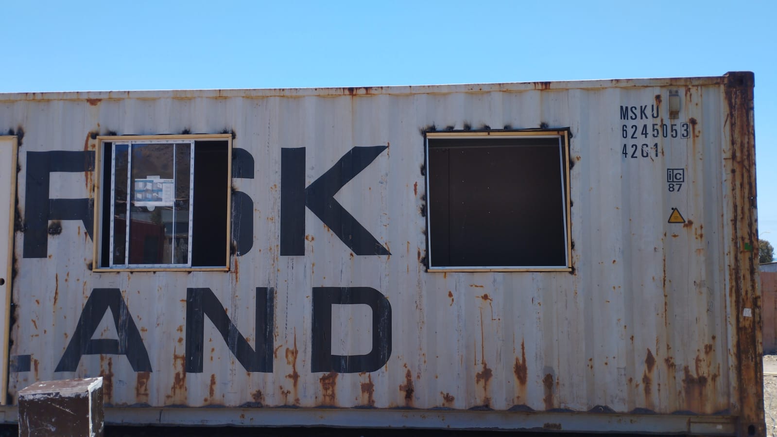 Contenedor blanco con el código MSKU 6245053 y dos aberturas para ventanas, una de ellas con parte del vidrio dañado.