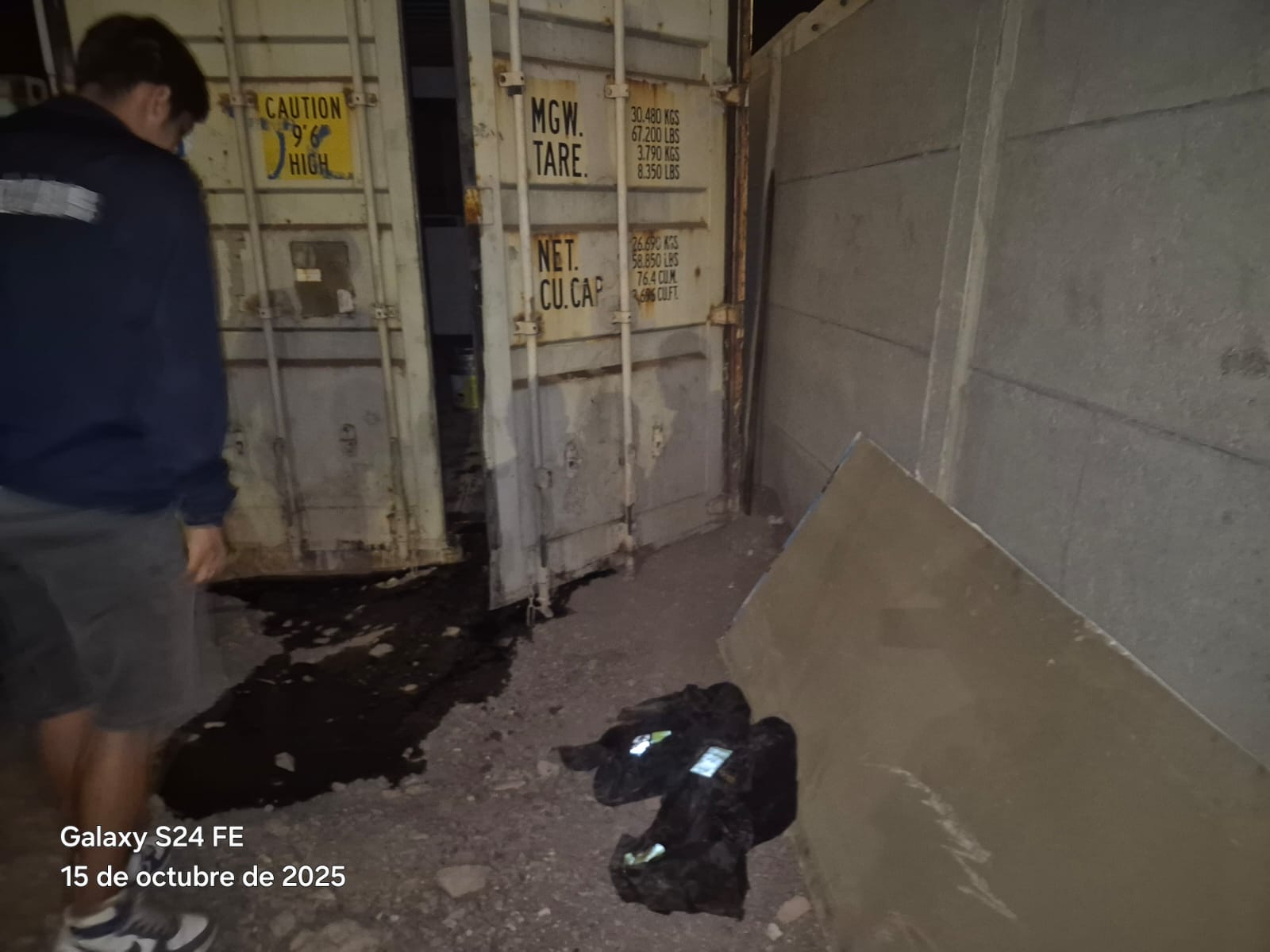 Interior oscuro de un contenedor con un derrame de líquido oscuro en el suelo y un hombre inspeccionando los daños.