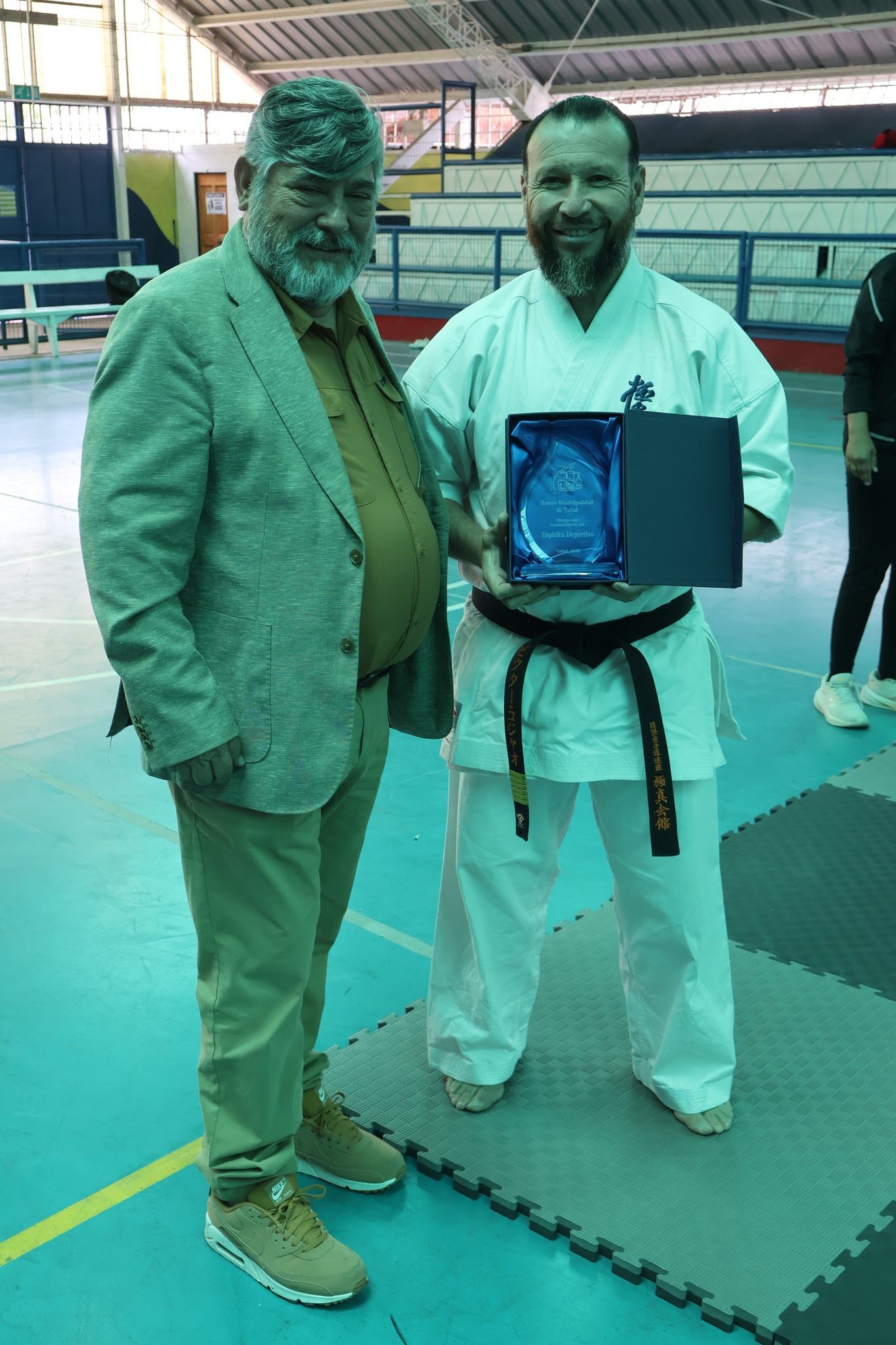 El instructor y campeón mundial de karate Víctor Collao recibe un reconocimiento municipal y posa con el director DIDECO Juan Mediano en el Polideportivo de Taltal.