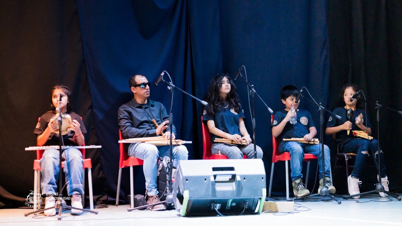 Ensamble musical con jóvenes y adultos tocando instrumentos de viento andino o percusión sentados en el escenario.