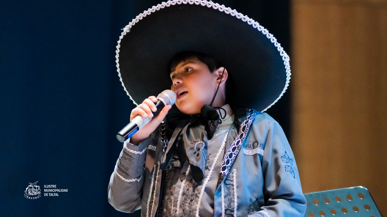 Primer plano de un niño solista del Taller de Canto con un traje de mariachi y sombrero grande, cantando al micrófono.
