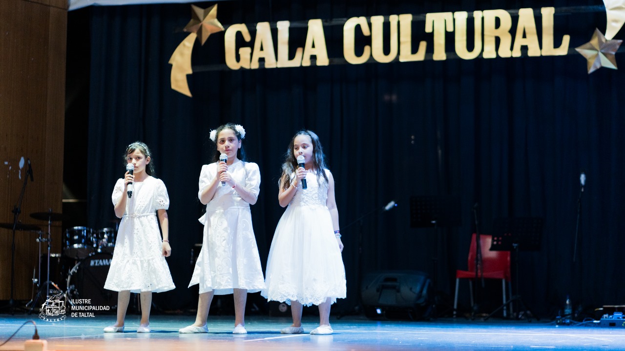 Trío de niñas del Taller de Canto interpretando una canción con micrófonos en mano, vestidas de blanco en el escenario.