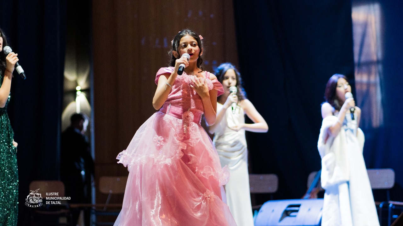 Niña solista en vestido de gala rosa, acompañada por otras cantantes con vestidos blancos.