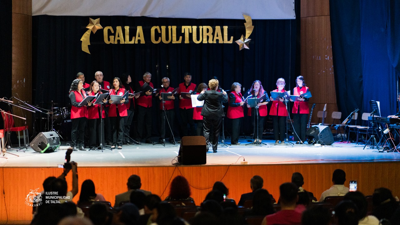 Coro de Cámara en chalecos rojos cantando y siendo dirigido en el escenario de la Gala Cultural, con público de fondo.