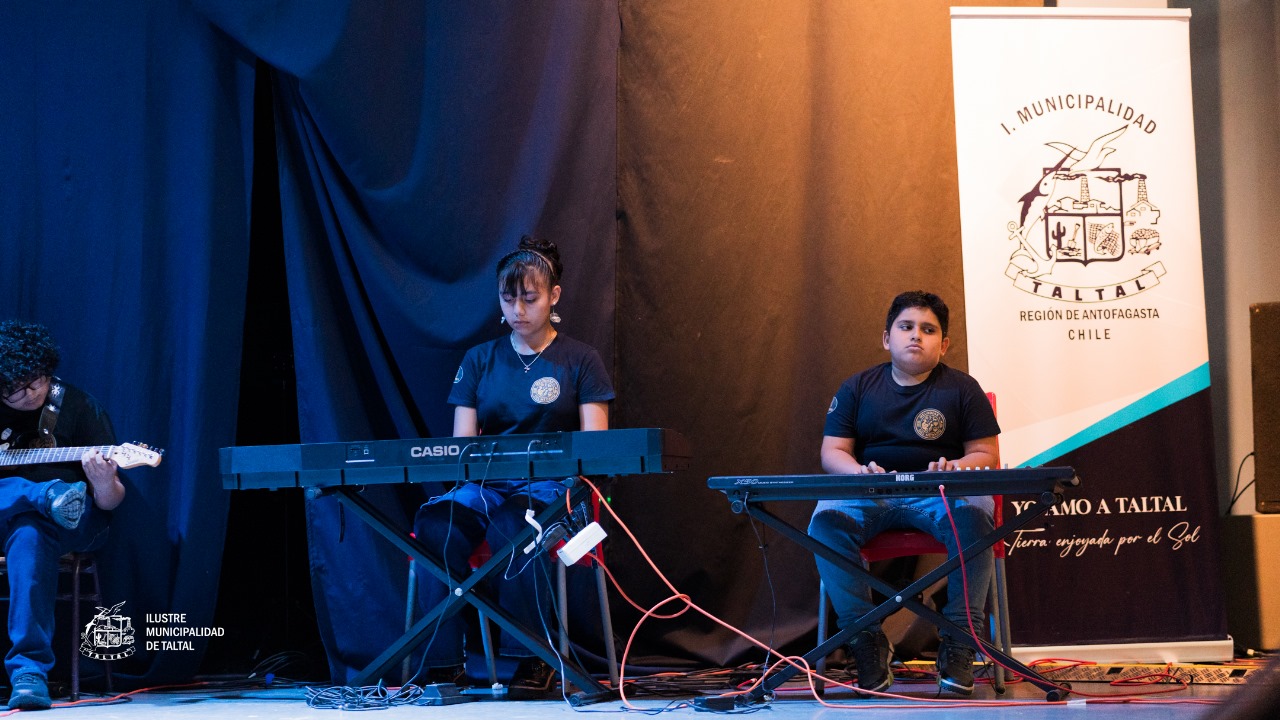 Tres estudiantes interpretando música con teclados y guitarra junto a un estandarte de la I. Municipalidad de Taltal.