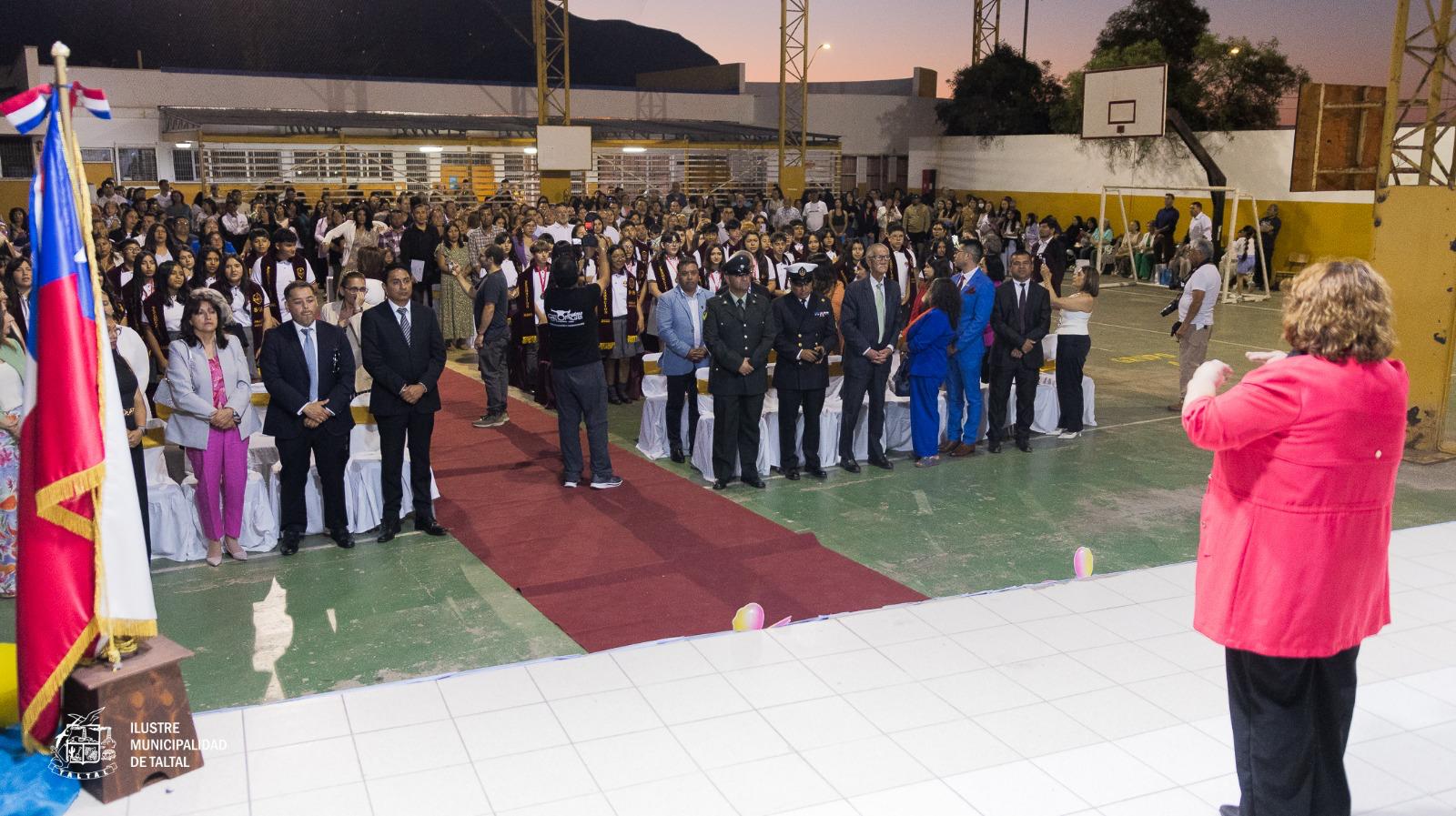 Autoridades locales y público de pie frente al escenario durante una ceremonia escolar.