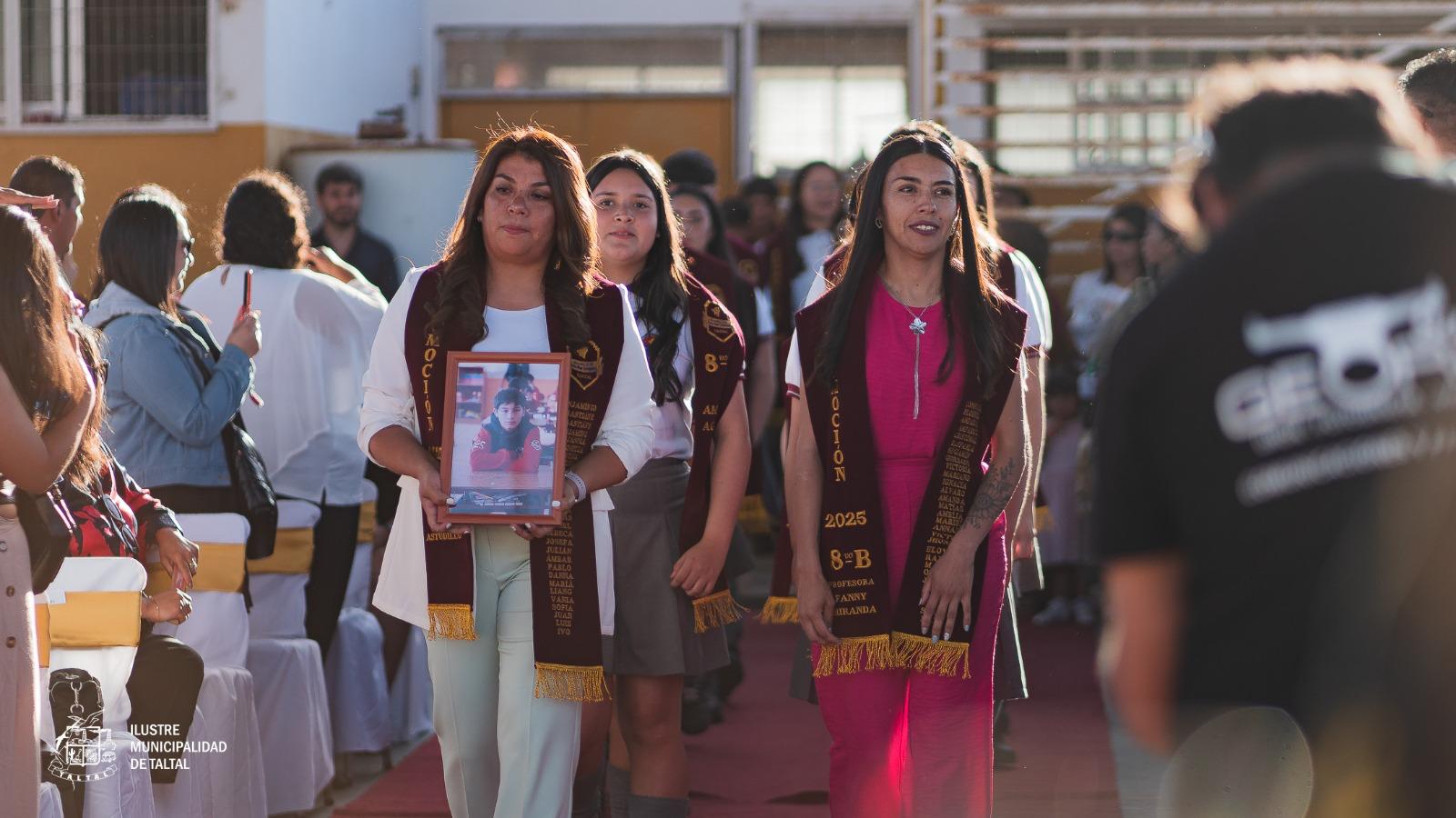 Dos docentes lideran el ingreso de los alumnos licenciados por la alfombra roja, una de ellas portando un cuadro conmemorativo.