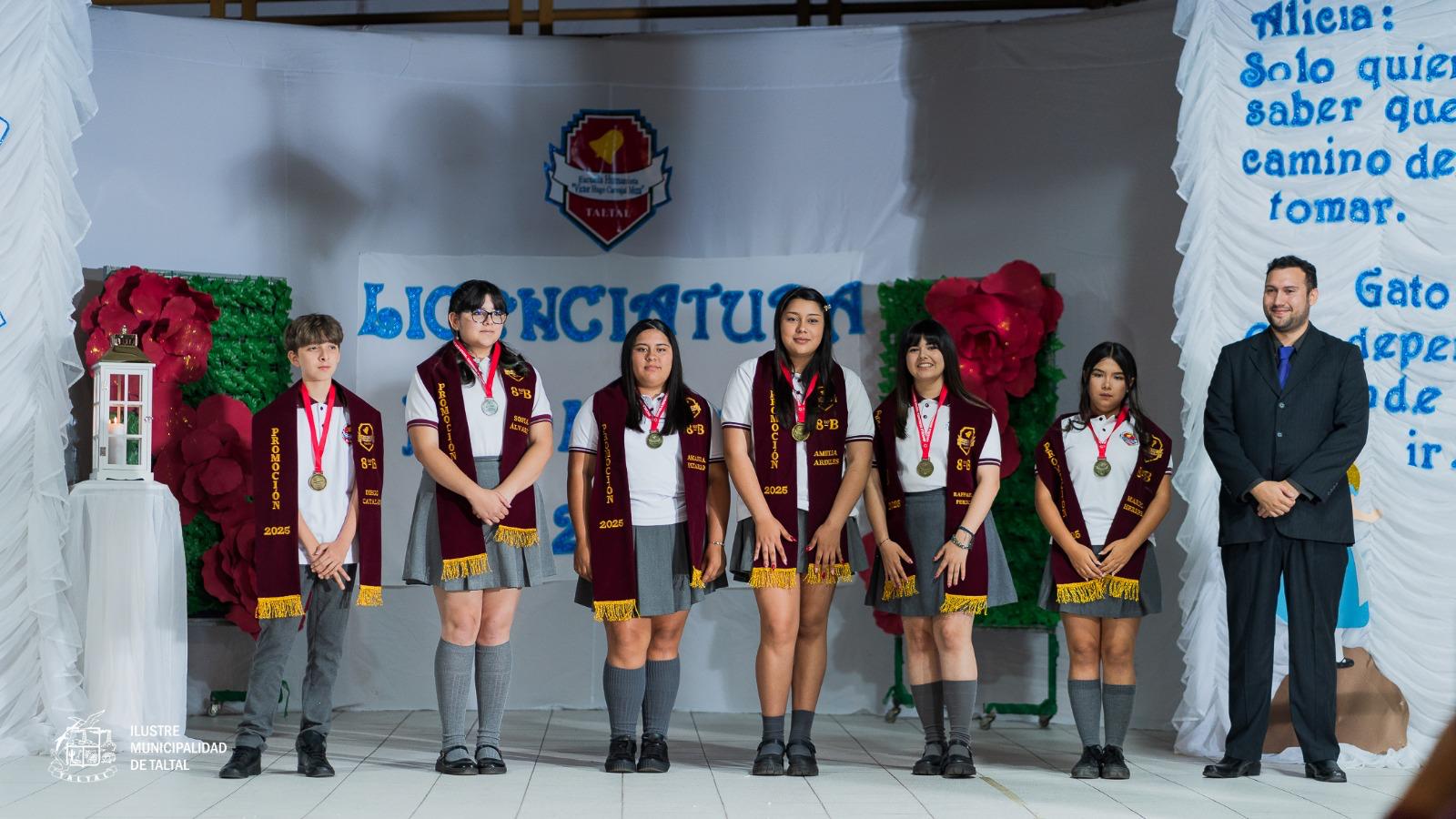 Grupo de estudiantes con estolas de graduación posando en el escenario junto a su profesor.