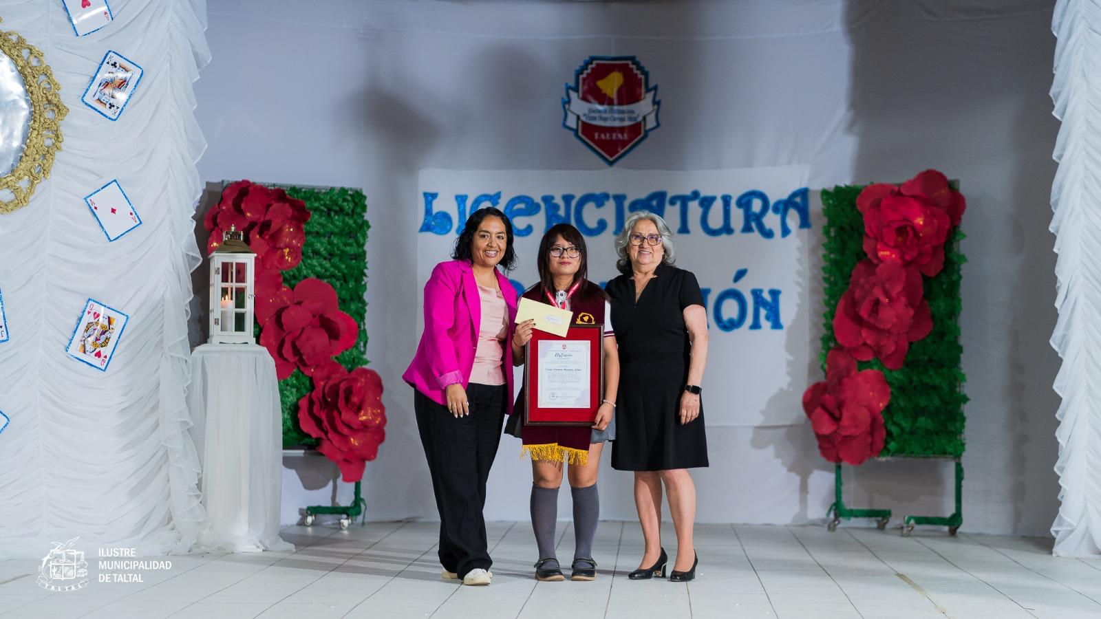 Una estudiante recibe su licencia de egreso acompañada por dos profesoras en el escenario.