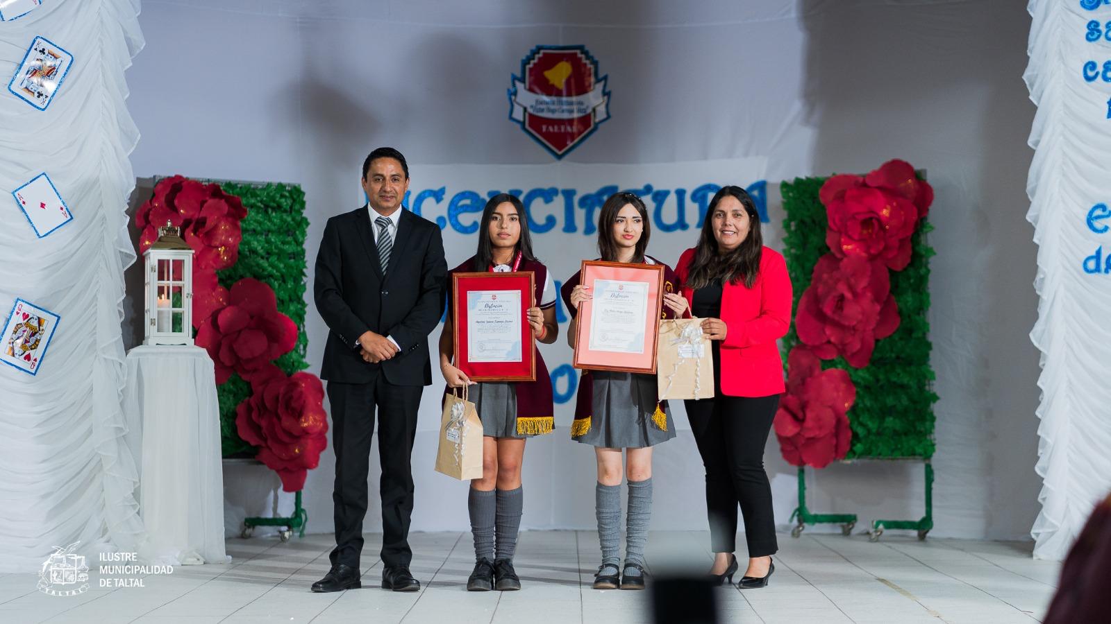 Dos alumnas egresadas posan con sus diplomas junto a autoridades académicas en el escenario.