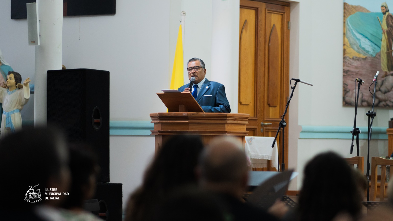 Hombre con traje hablando desde el ambón de la Parroquia San Francisco Javier durante el evento del Coro de Cámara.