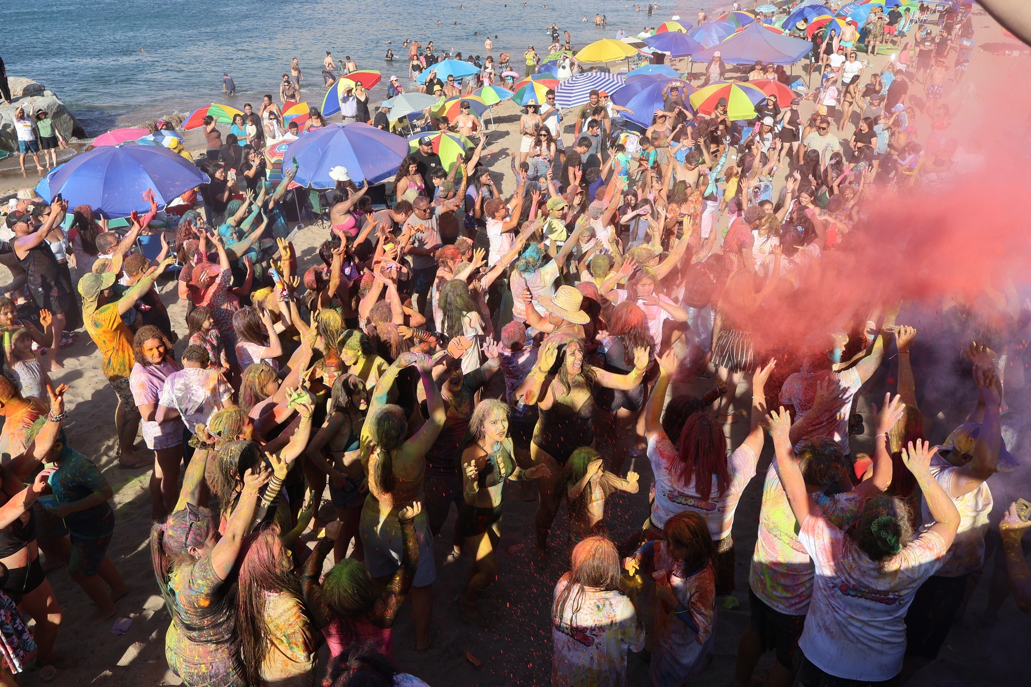 Jóvenes celebrando con las manos en alto bajo una lluvia de polvos de colores en la playa.