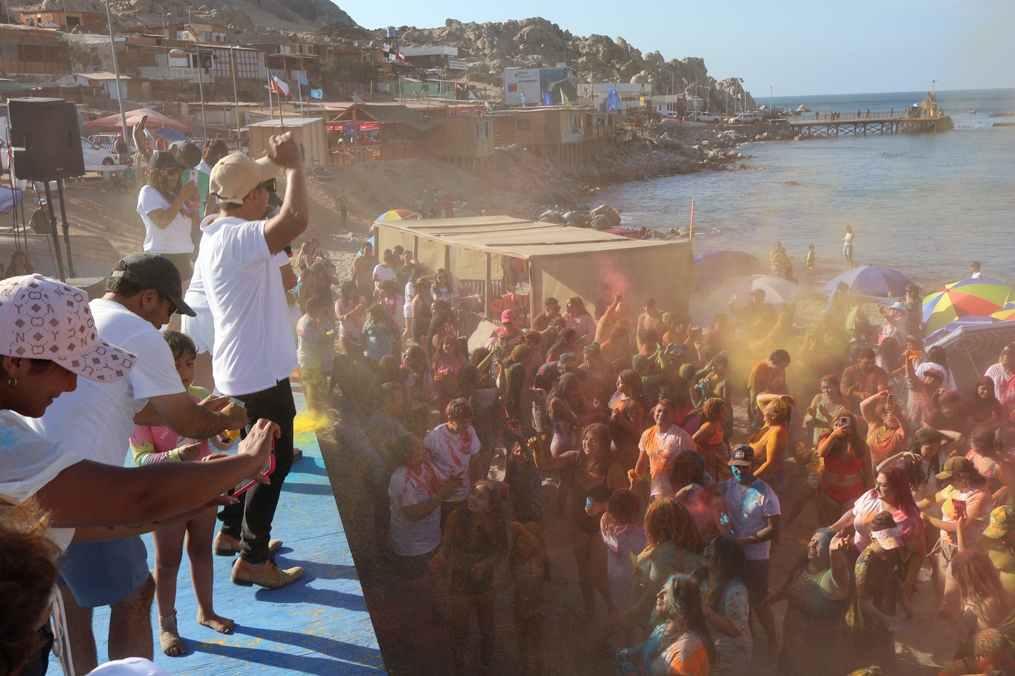 Nube de polvos amarillos y naranjas lanzada desde el escenario hacia los asistentes en la playa.