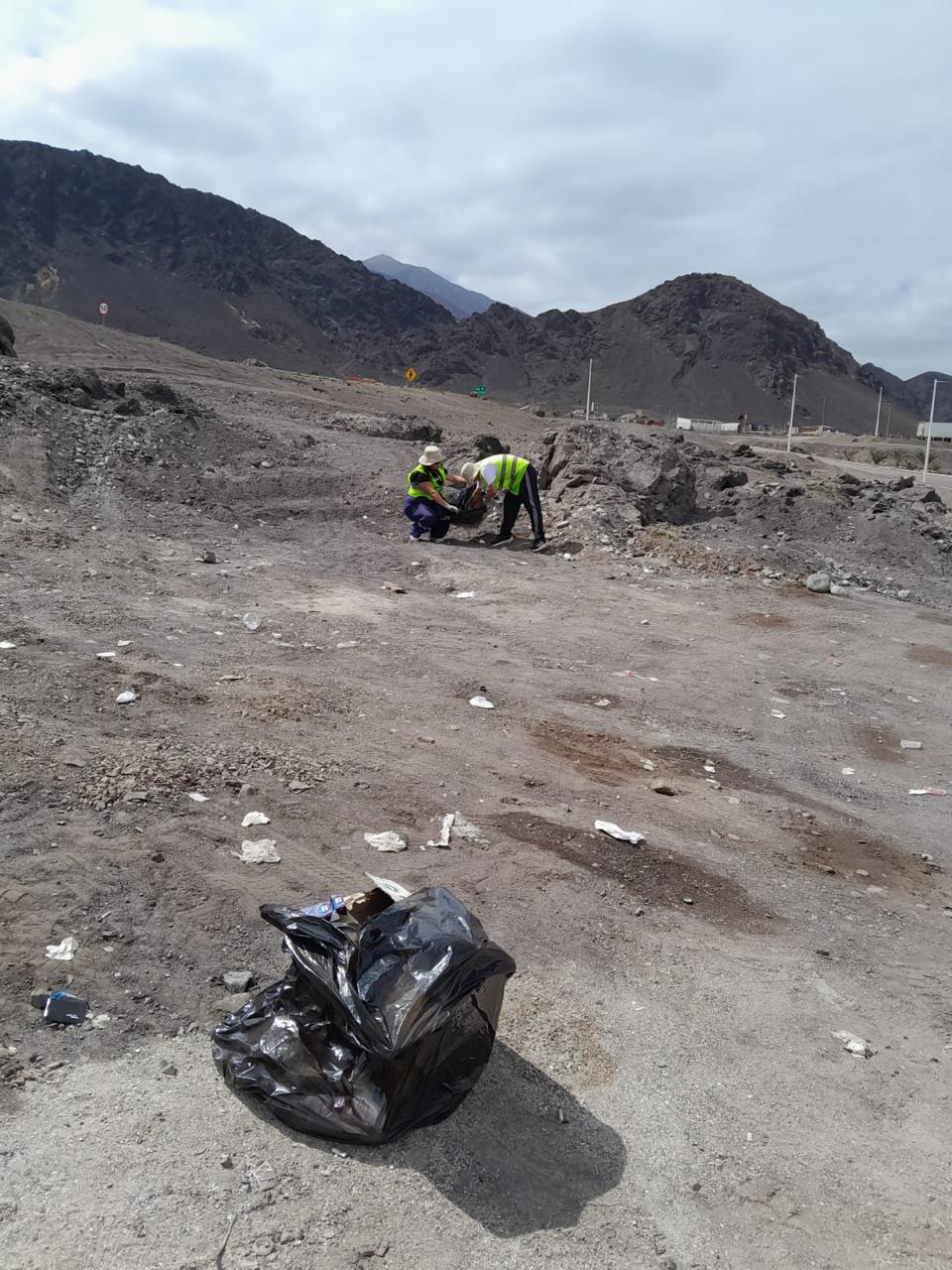 Participantes del Team Verano Taltal recolectan basura en terreno, con bolsa de residuos lista para retiro en primer plano.