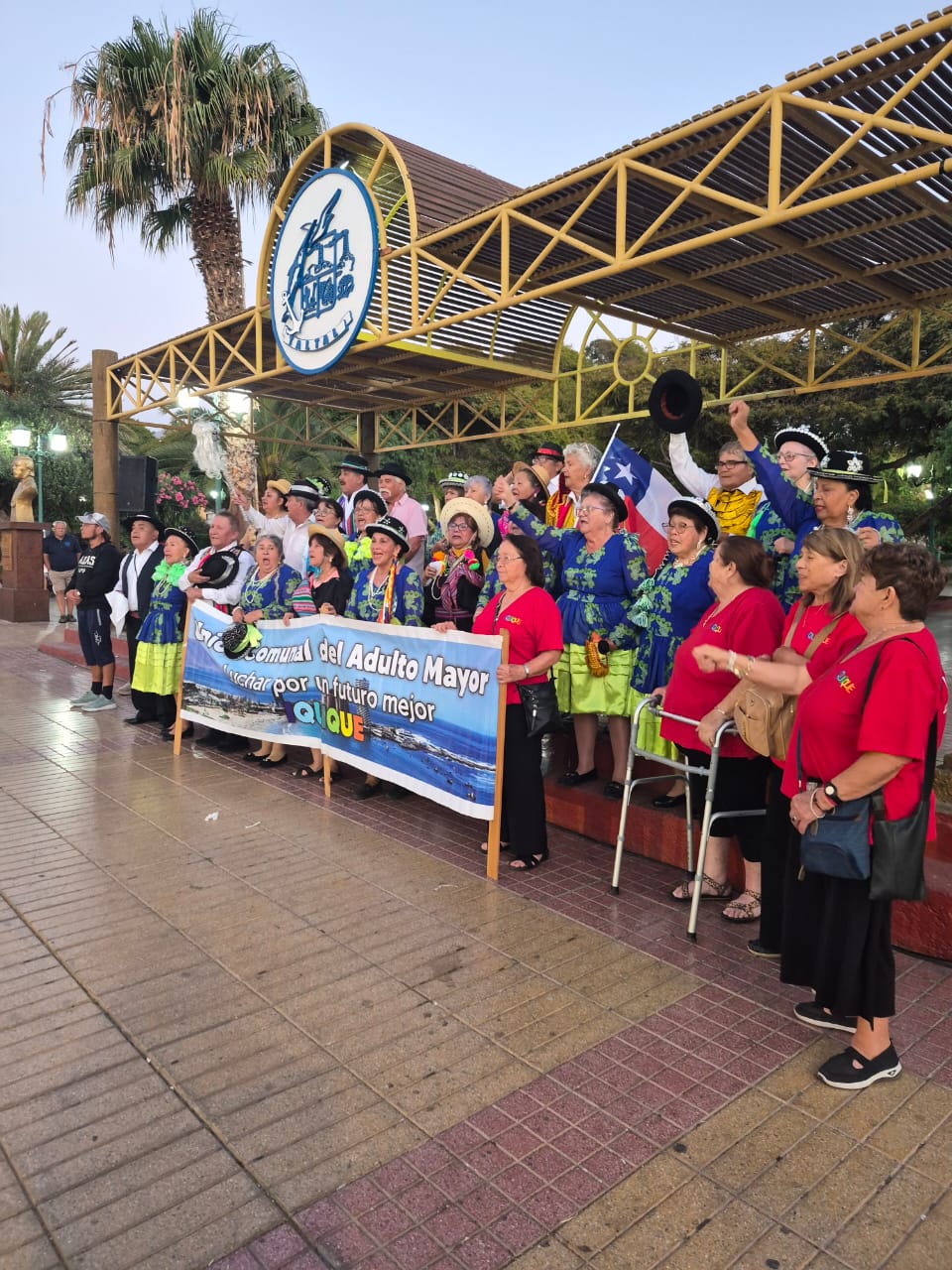 Grupo de adultos mayores con vestimenta folclórica y lienzo institucional se reúne en la Plaza de Armas de Taltal, con bandera de Chile al fondo.
