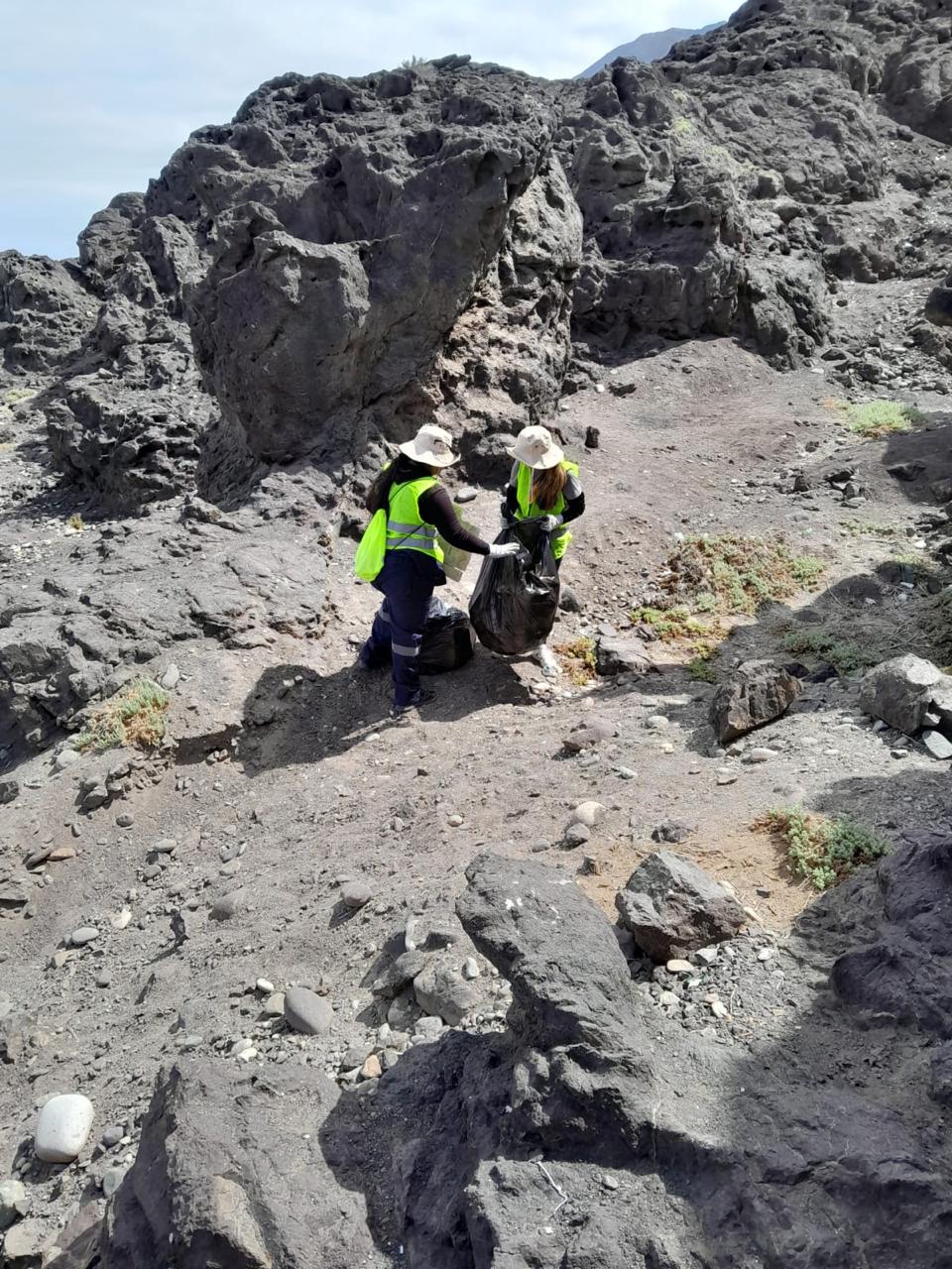 Dos integrantes del Team Verano Taltal juntan residuos y cierran bolsa de basura durante limpieza en zona de roqueríos.