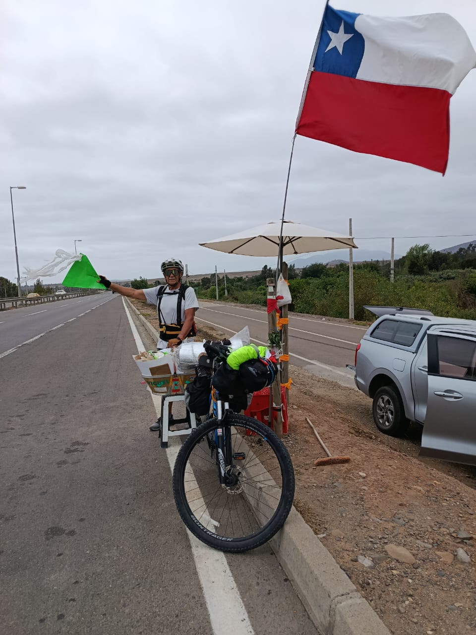 Pedro Lemus junto a una bandera de Chile instalada al costado de la ruta, en una parada de apoyo durante su travesía en bicicleta.