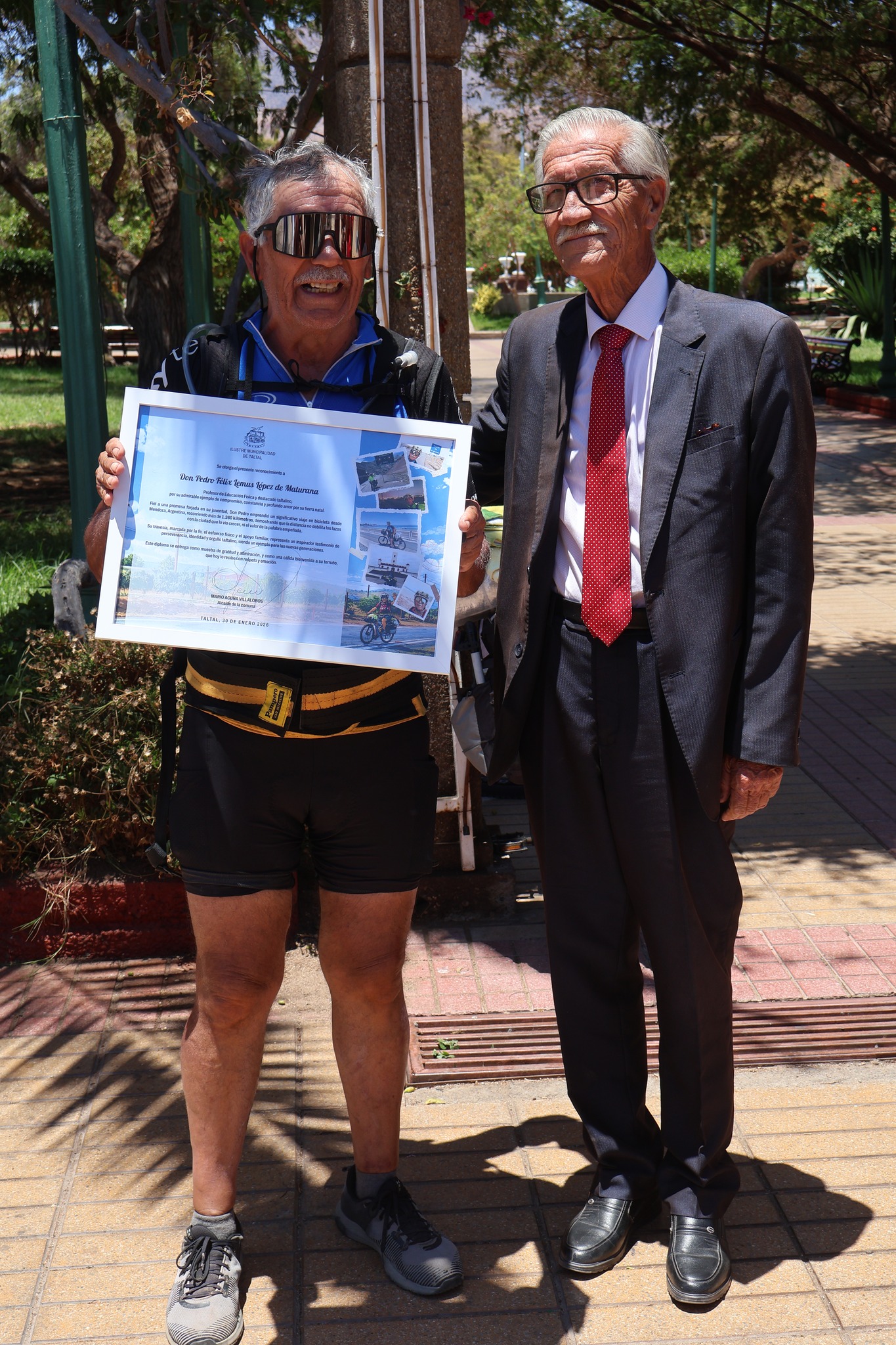 Pedro Lemus posa en la plaza de Taltal junto a una autoridad, sosteniendo un reconocimiento por su travesía en bicicleta desde Mendoza.