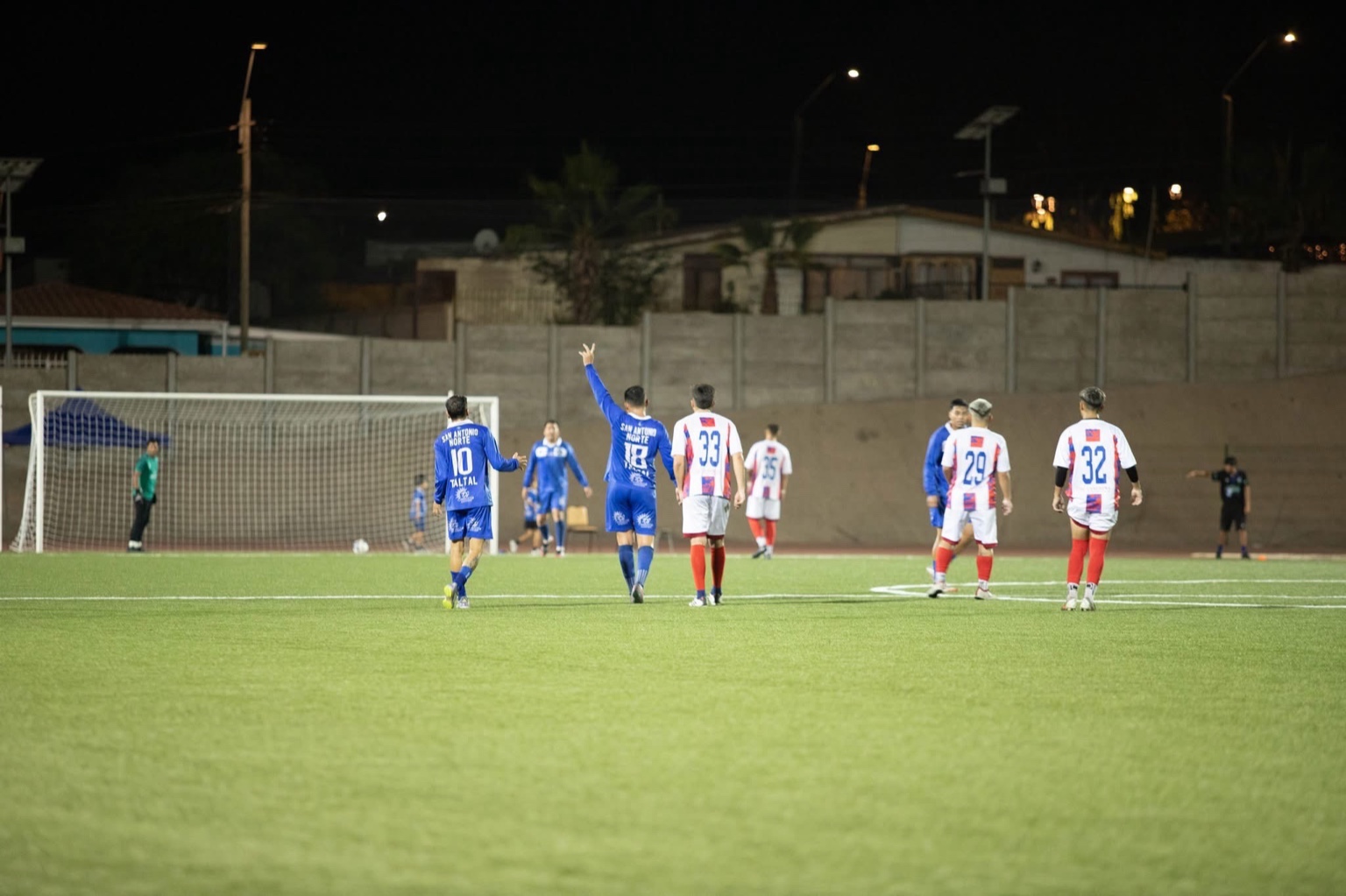 Jugadores de San Antonio Norte de Taltal caminando en la cancha tras una jugada, con el estadio y muros de fondo de noche.
