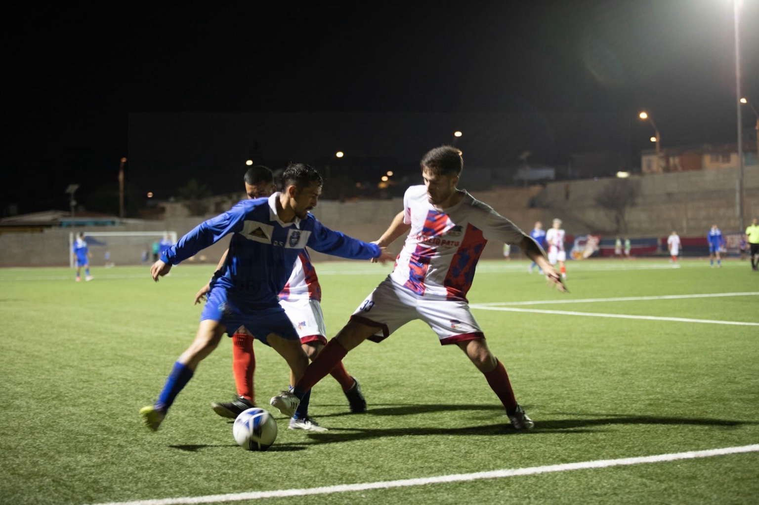 Jugada del partido con disputa de balón entre jugadores de San Antonio Norte y rival, en acción nocturna sobre césped sintético.